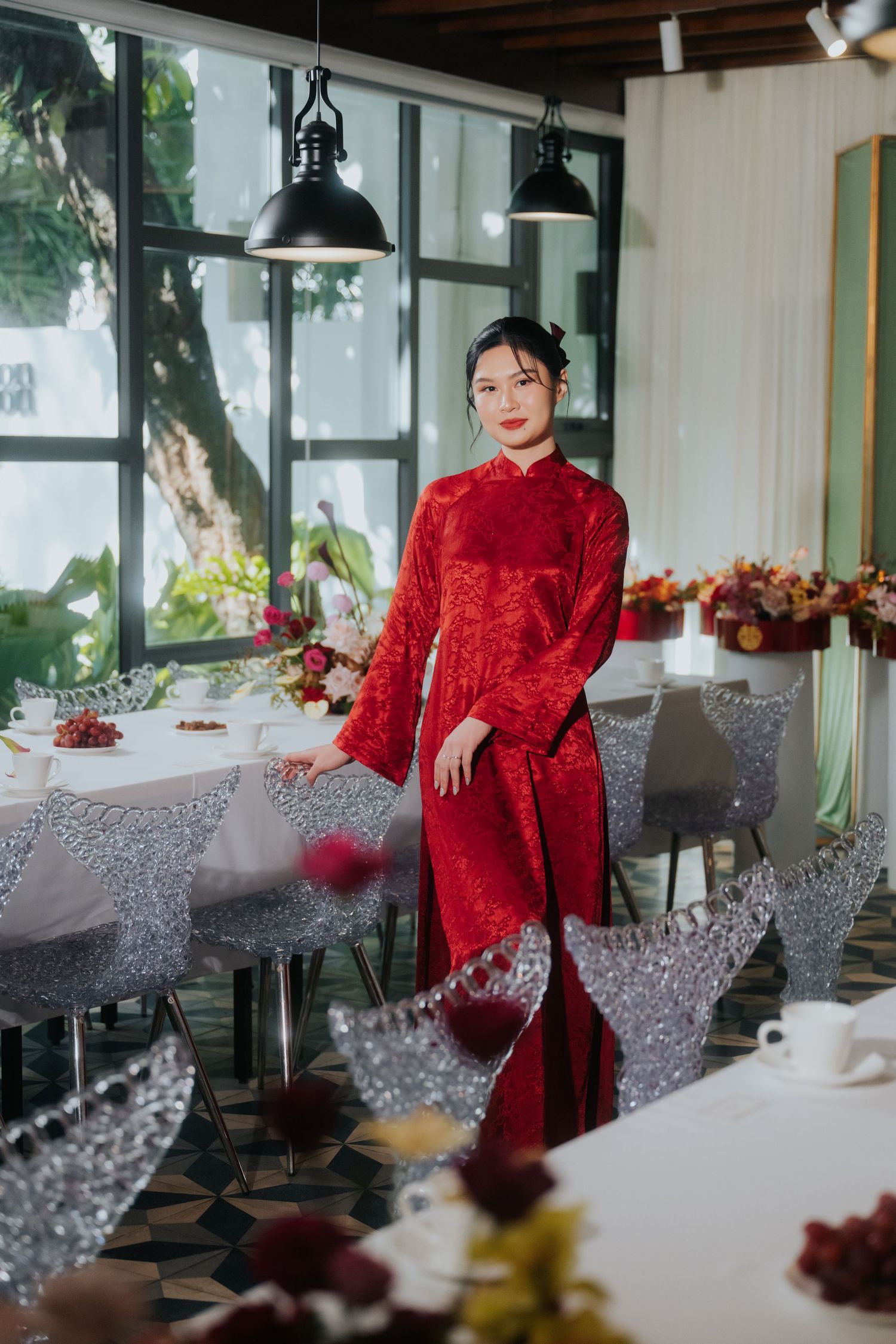 Woman in red lace ao dai dress standing elegantly in a decorated restaurant with crystal chairs and floral arrangements.
