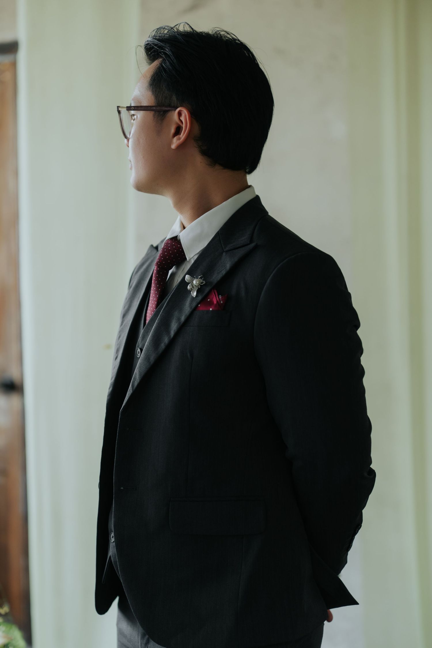 Man in black suit with red tie and pocket square, wearing glasses, standing sideways in elegant formal attire.