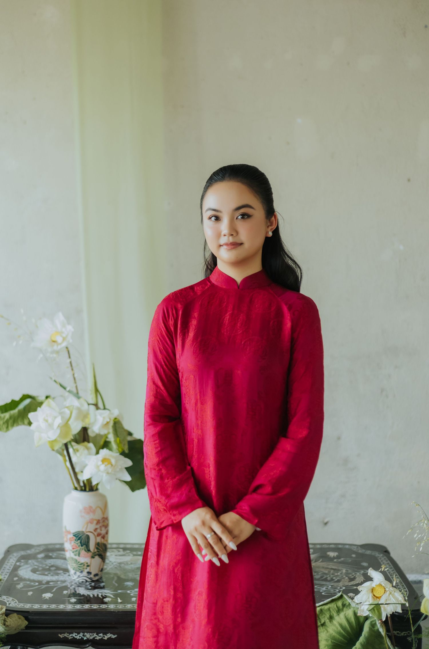 A young woman in a red áo dài traditional Vietnamese dress stands gracefully beside a vase of white lotus flowers.