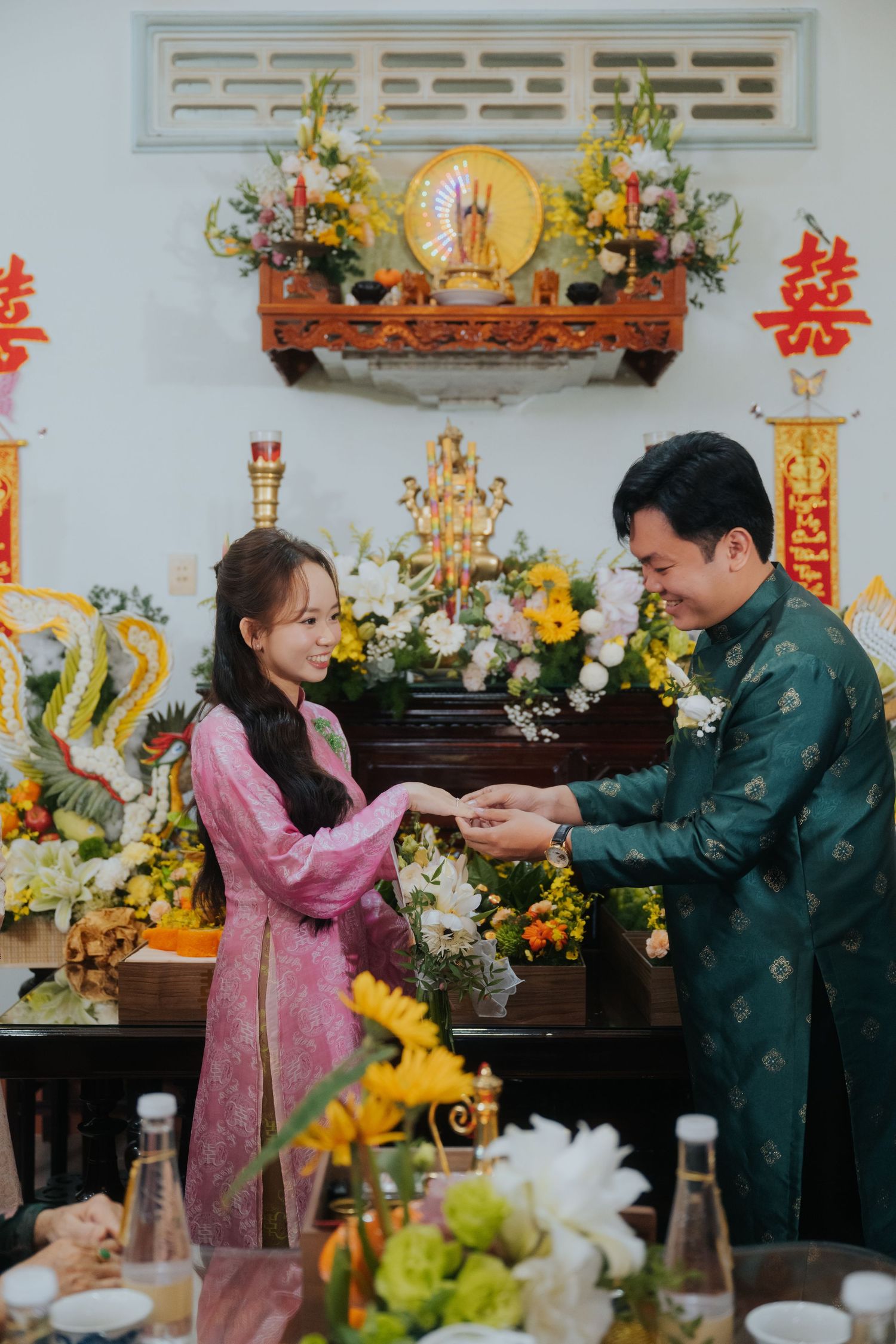 A couple in traditional Vietnamese ao dai attire exchange rings at a beautifully decorated altar during a traditional engagement ceremony.