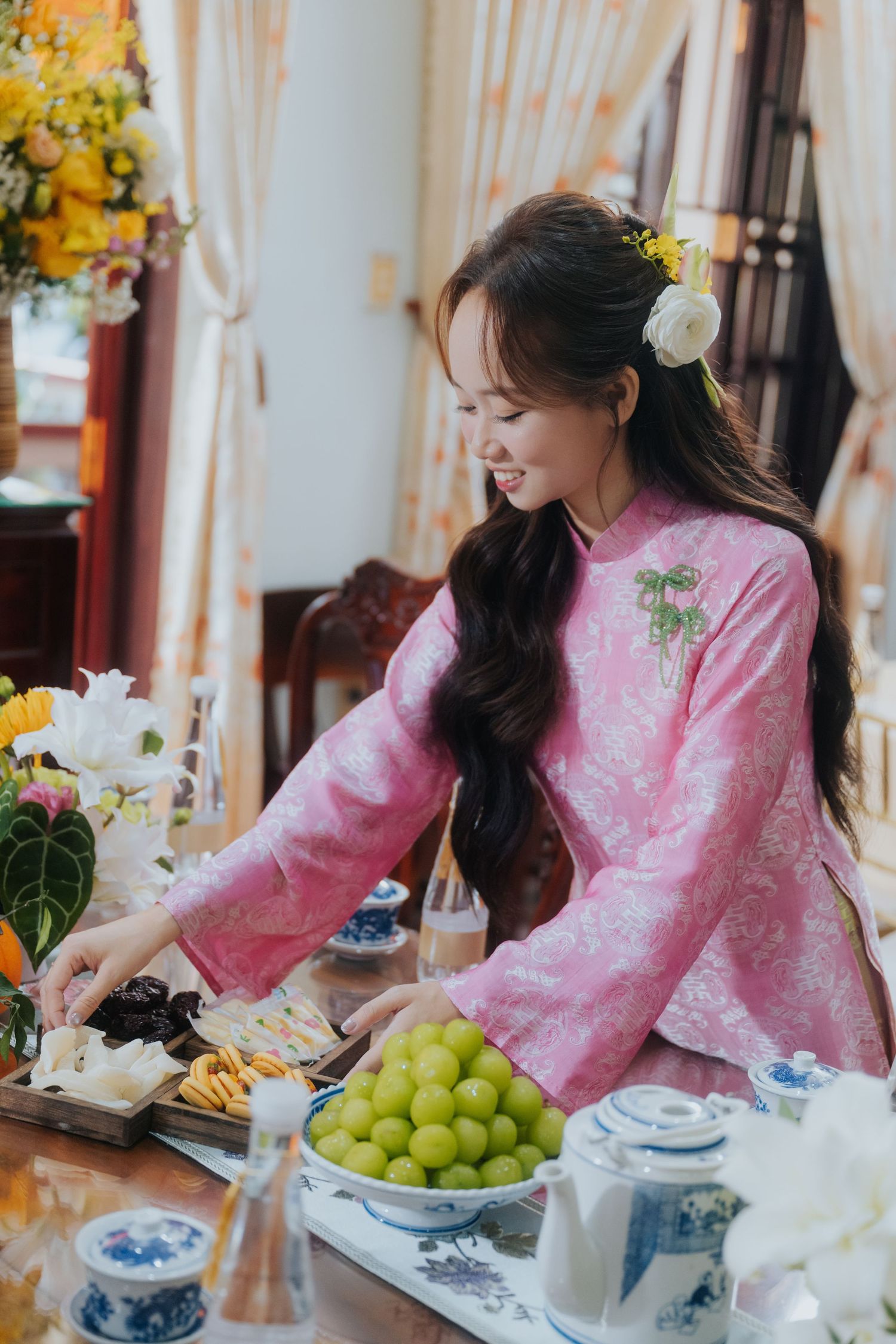 Young Vietnamese woman in pink ao dai arranges traditional food and green grapes on table during Tet celebration.