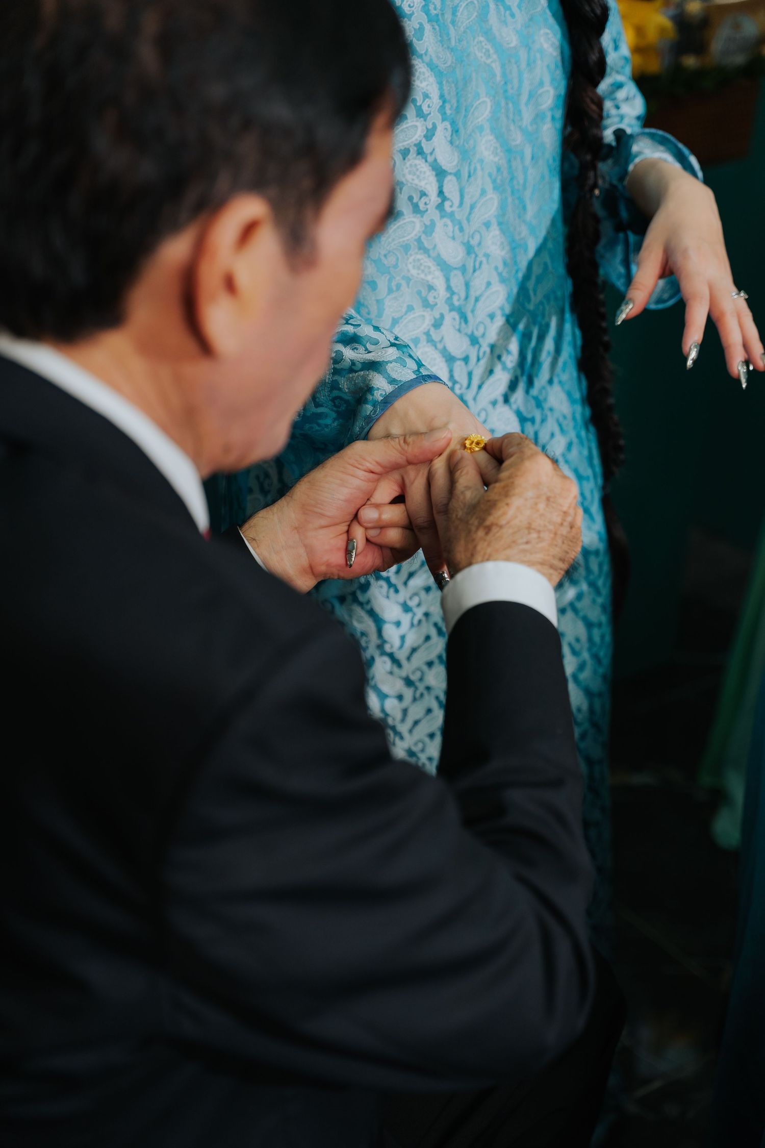 Man in black suit placing a gold ring on a woman's finger in a blue lace dress during a ceremony.