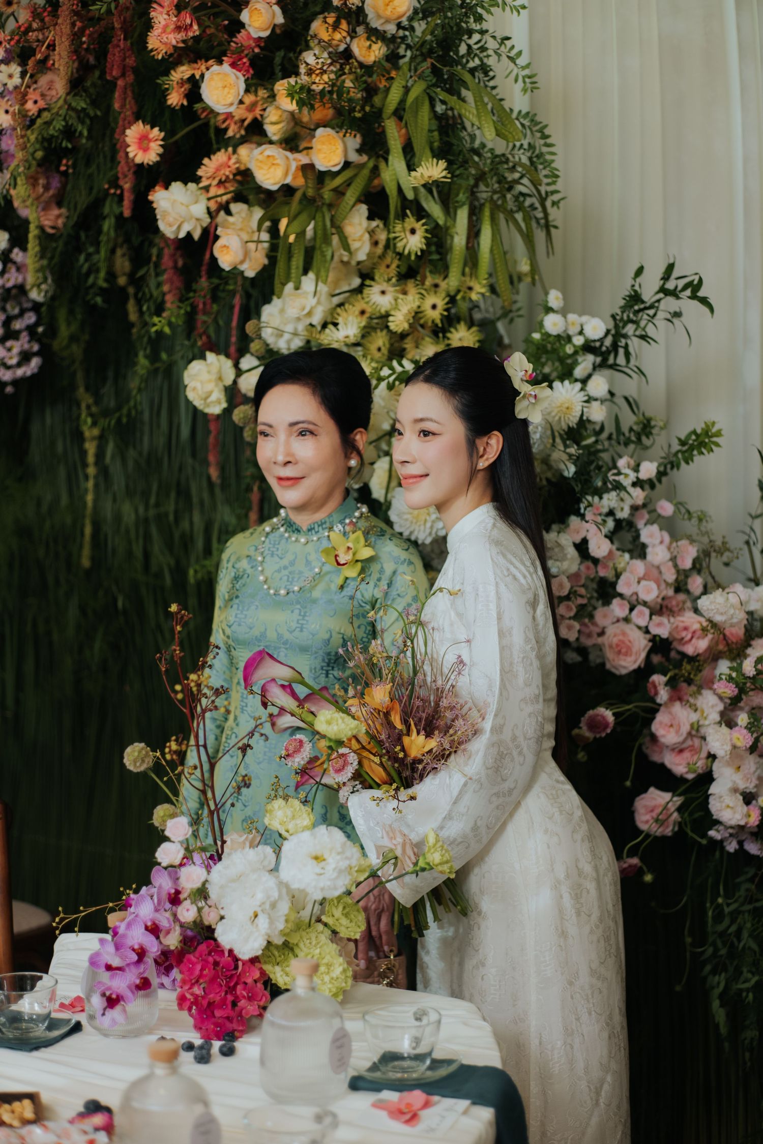 Two women in elegant ao dai attire pose together at a floral wedding ceremony backdrop with pink and yellow blooms.