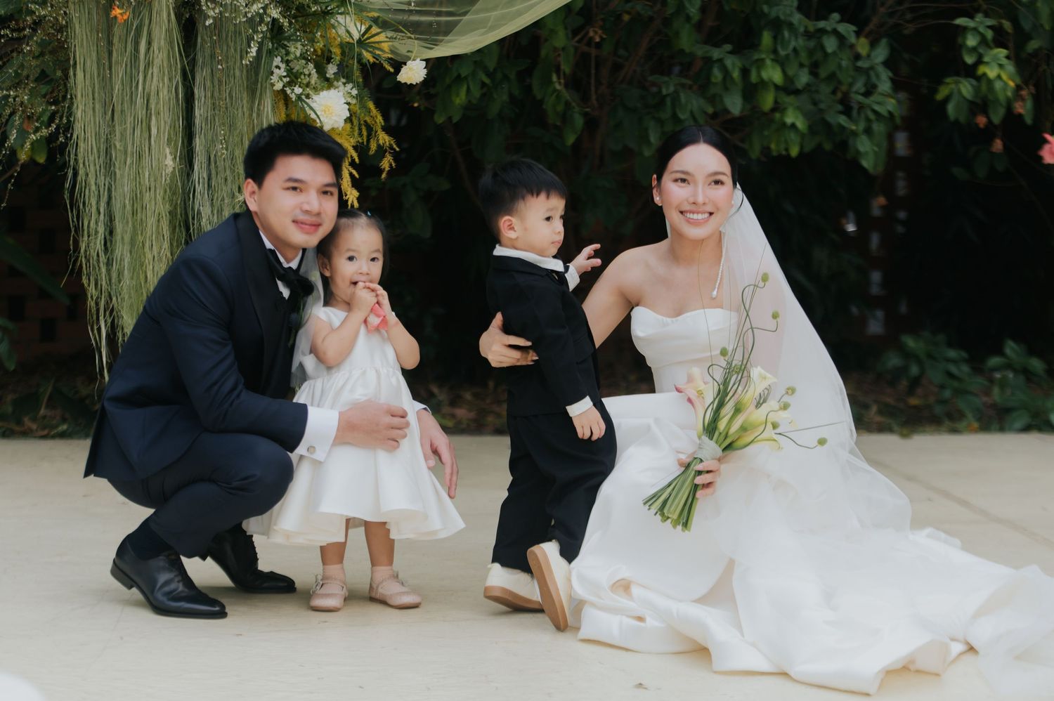 Asian couple in wedding attire crouching with two young children dressed formally, posing outdoors at a garden wedding ceremony.