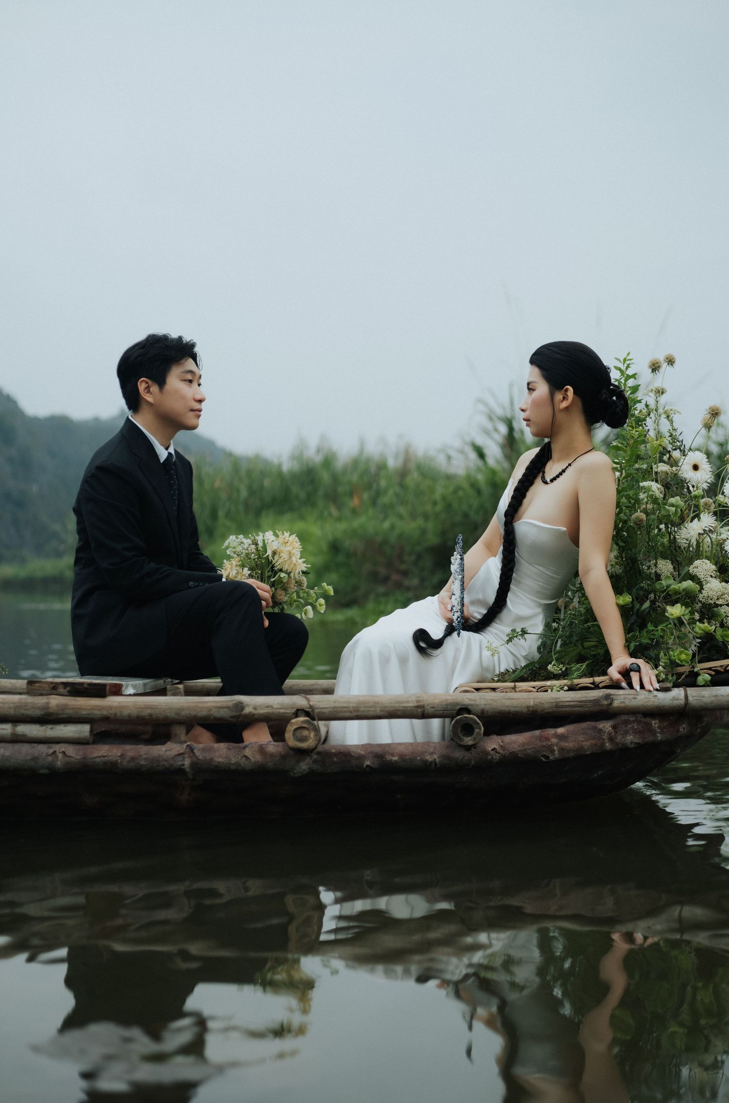 A couple in formal attire sits on a wooden boat surrounded by flowers on a calm river with lush greenery.