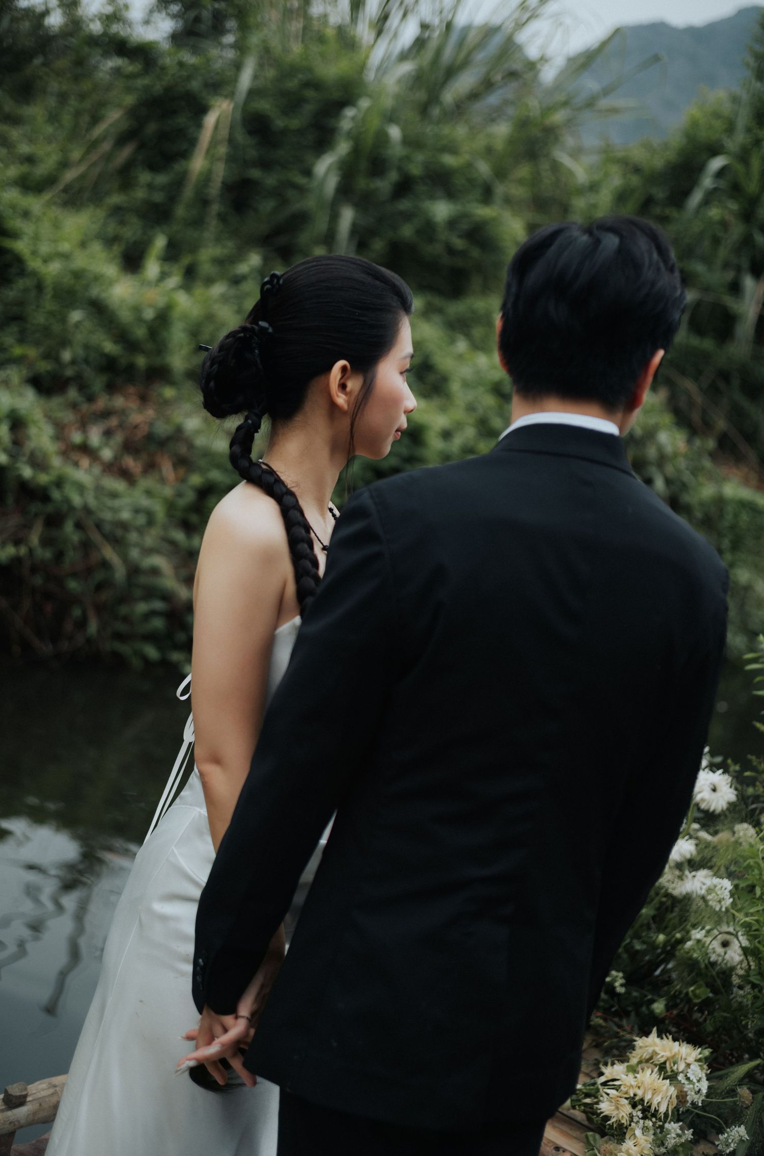 A bride and groom hold hands facing a lush green river backdrop during an outdoor wedding ceremony.