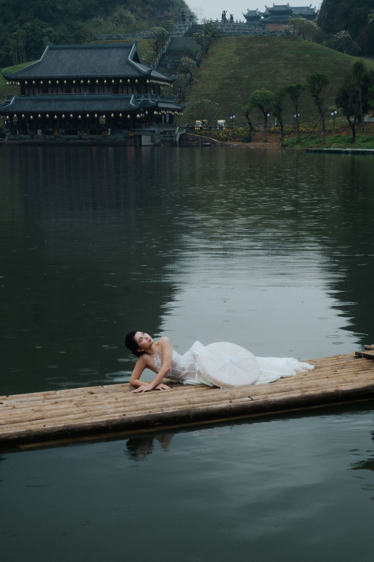 Woman in white dress reclining on bamboo raft over calm lake with traditional Asian temple and lush hills in background.