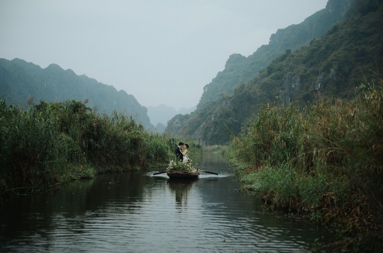 A person rowing a small boat through a misty river surrounded by lush greenery and dramatic karst mountains in Vietnam.