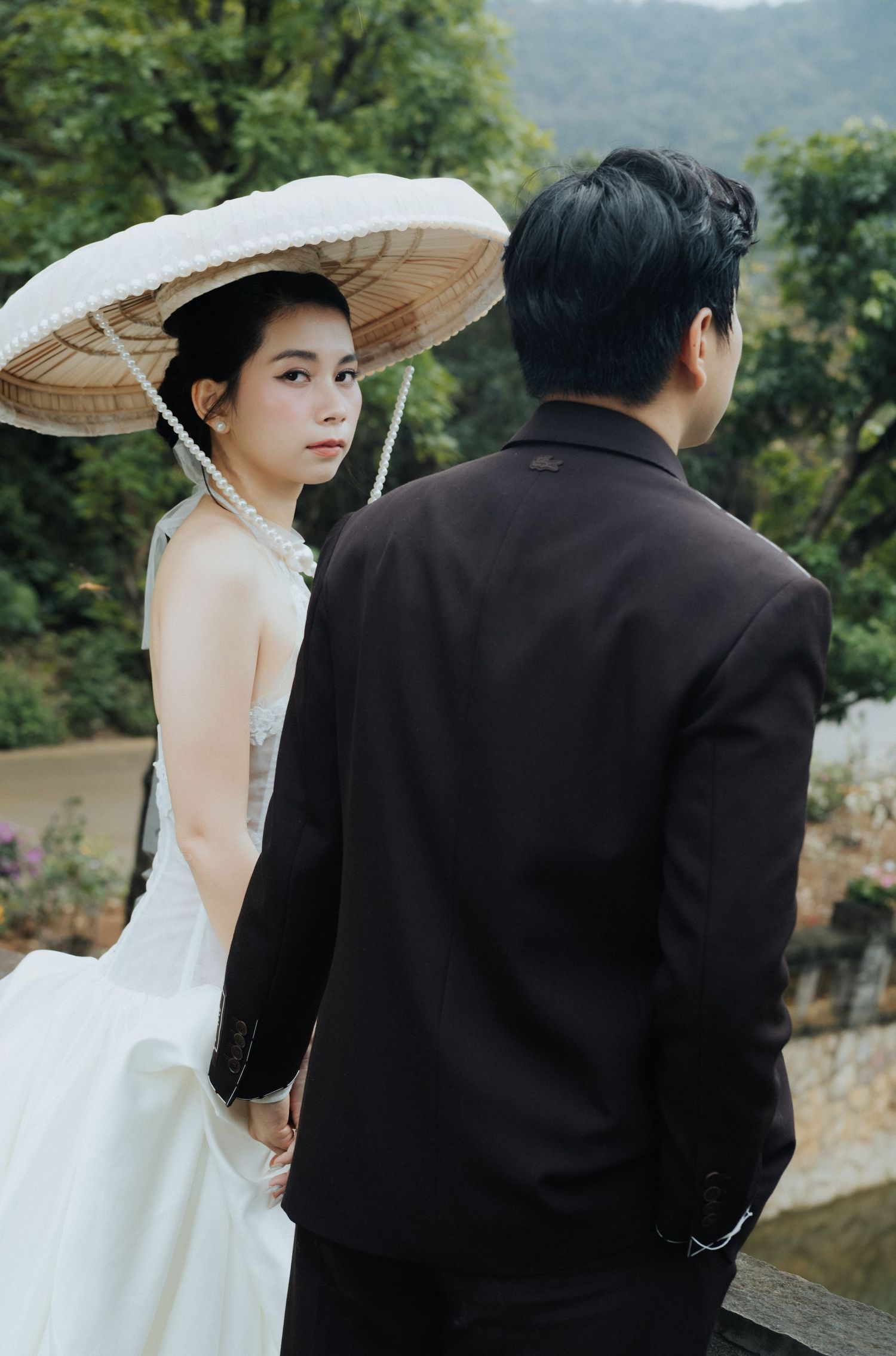 A bride in a white gown and traditional Vietnamese conical hat holds hands with a groom in a dark suit outdoors.