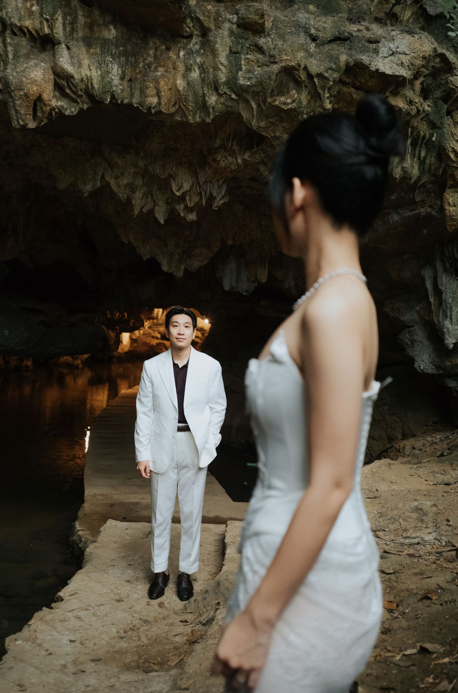 Couple in a cave setting, groom in white suit stands by water while bride in silver gown poses in foreground.