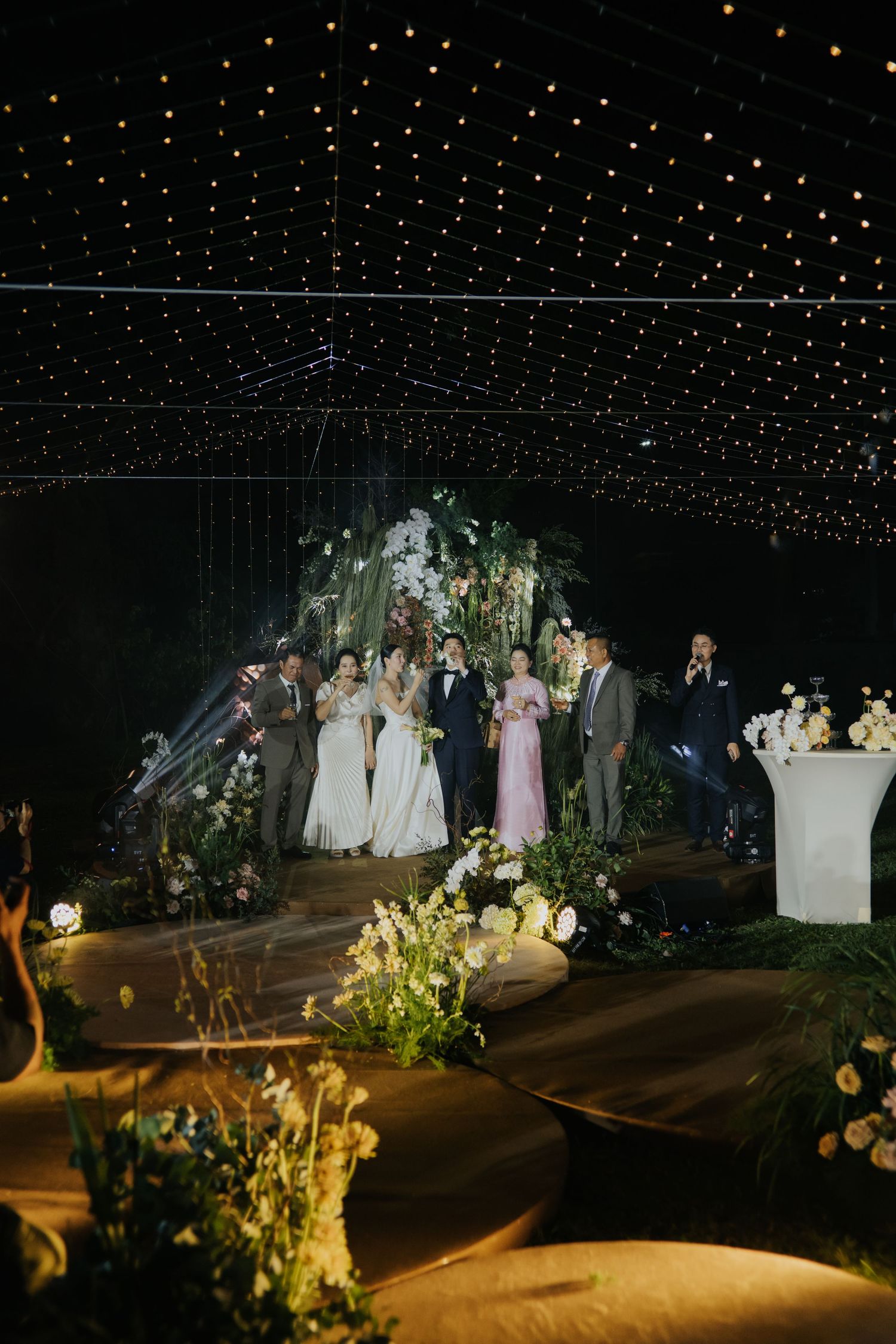 Elegant wedding reception under a canopy of twinkling fairy lights with bridal party on a floral-decorated stage.