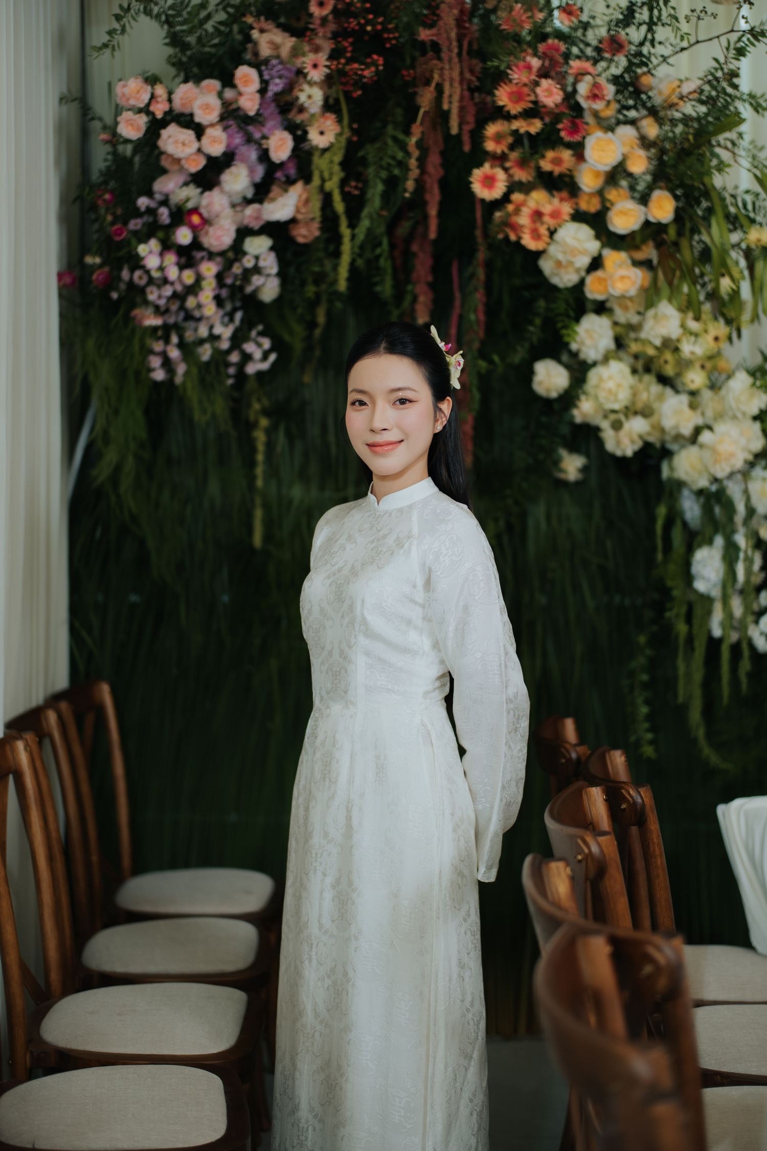 Woman in white ao dai dress stands before floral wall at elegant wedding venue.