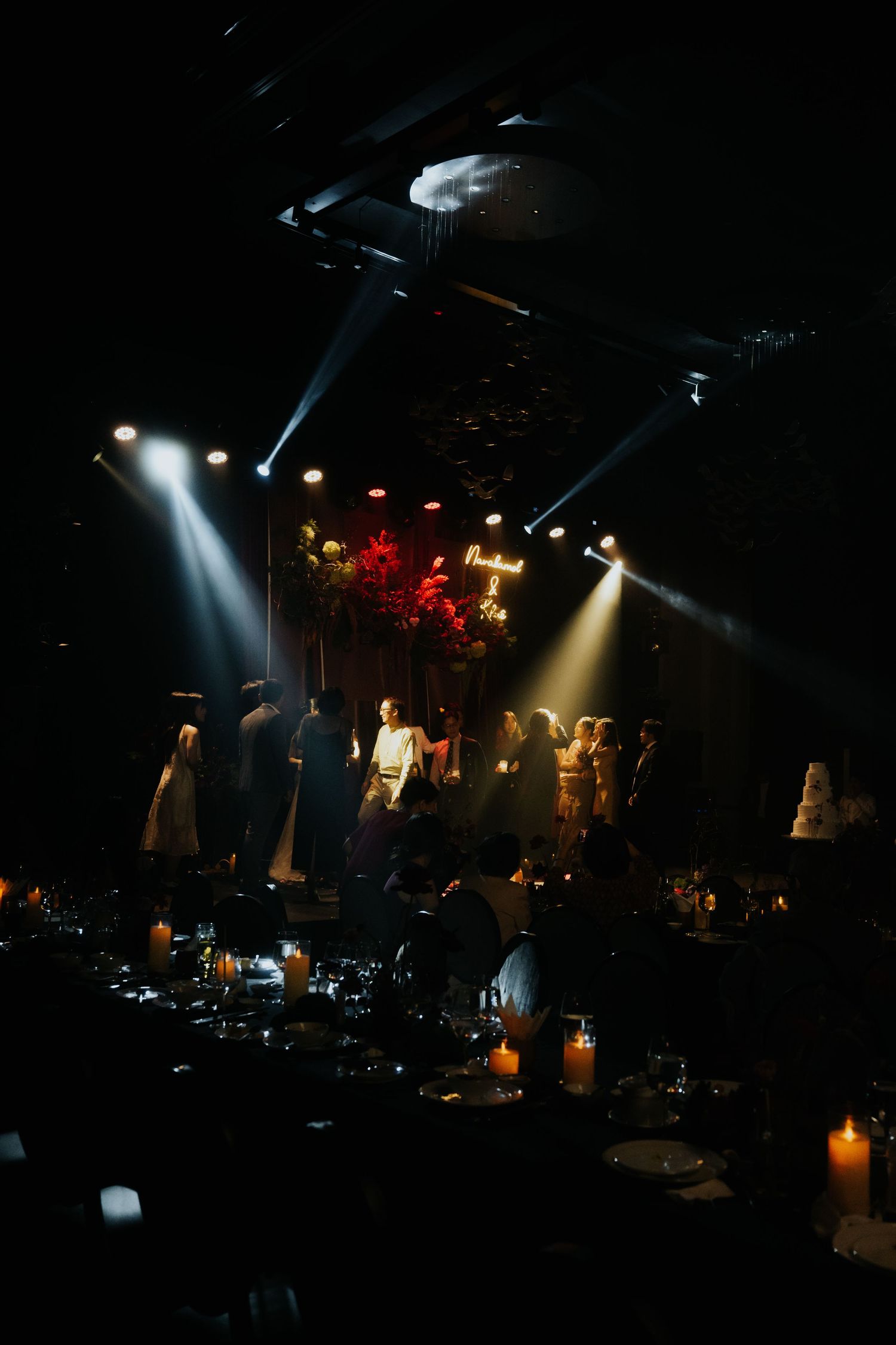 Elegant dark wedding reception venue with dramatic stage lighting, candles on tables, and guests gathered near a neon sign.