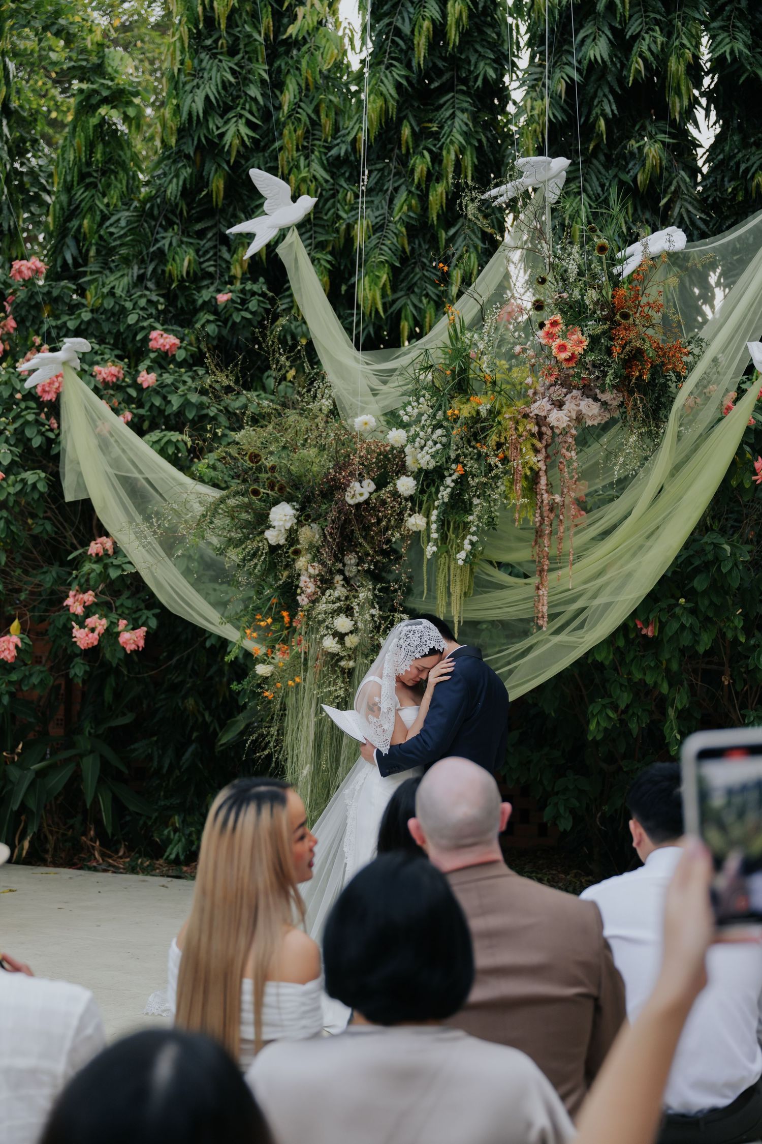 Couple kissing at outdoor wedding altar with green tulle, floral arrangements, and white doves, guests watching and photographing.