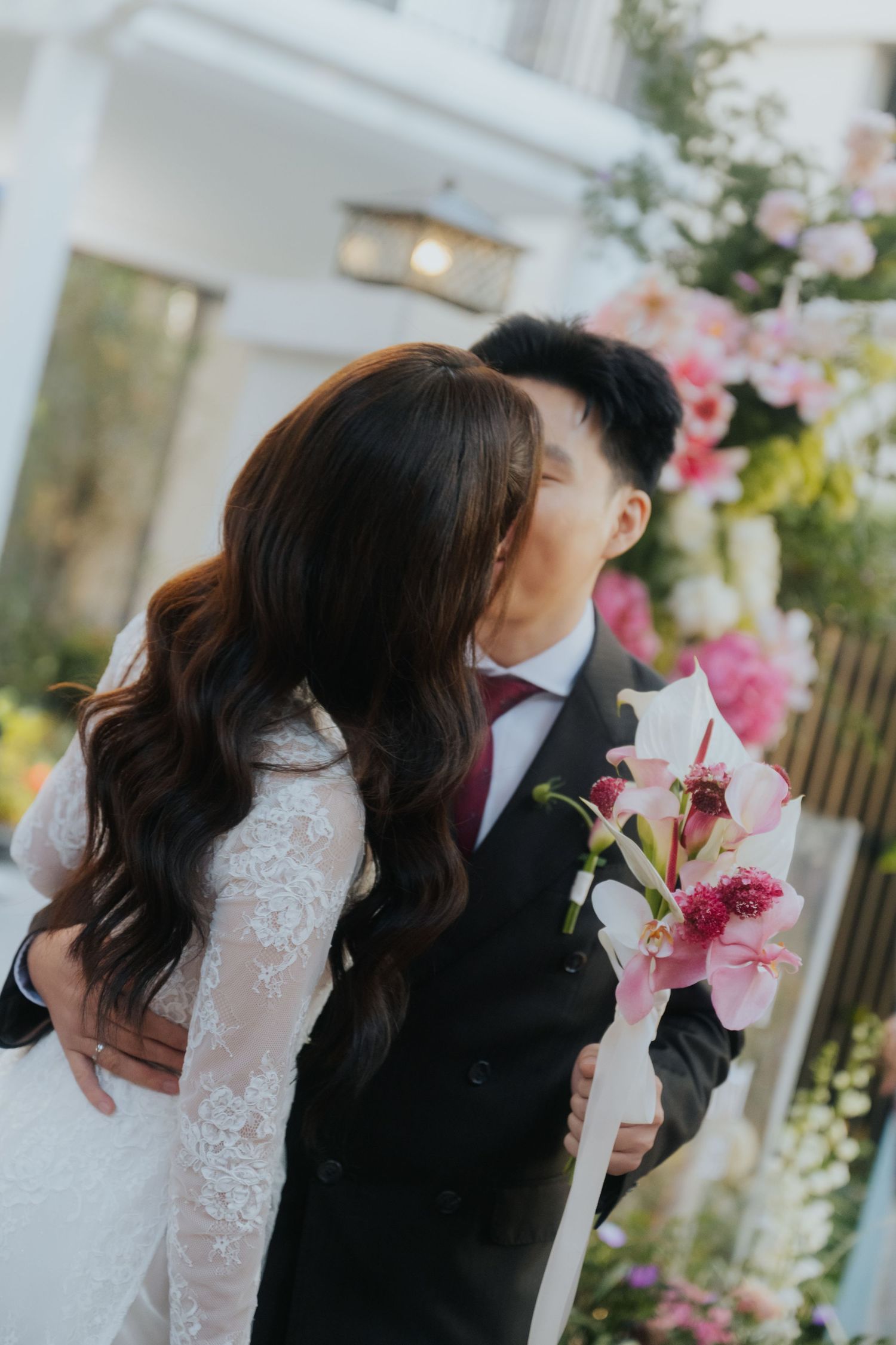 A bride and groom share a romantic kiss outdoors, with pink floral arrangements in the background.