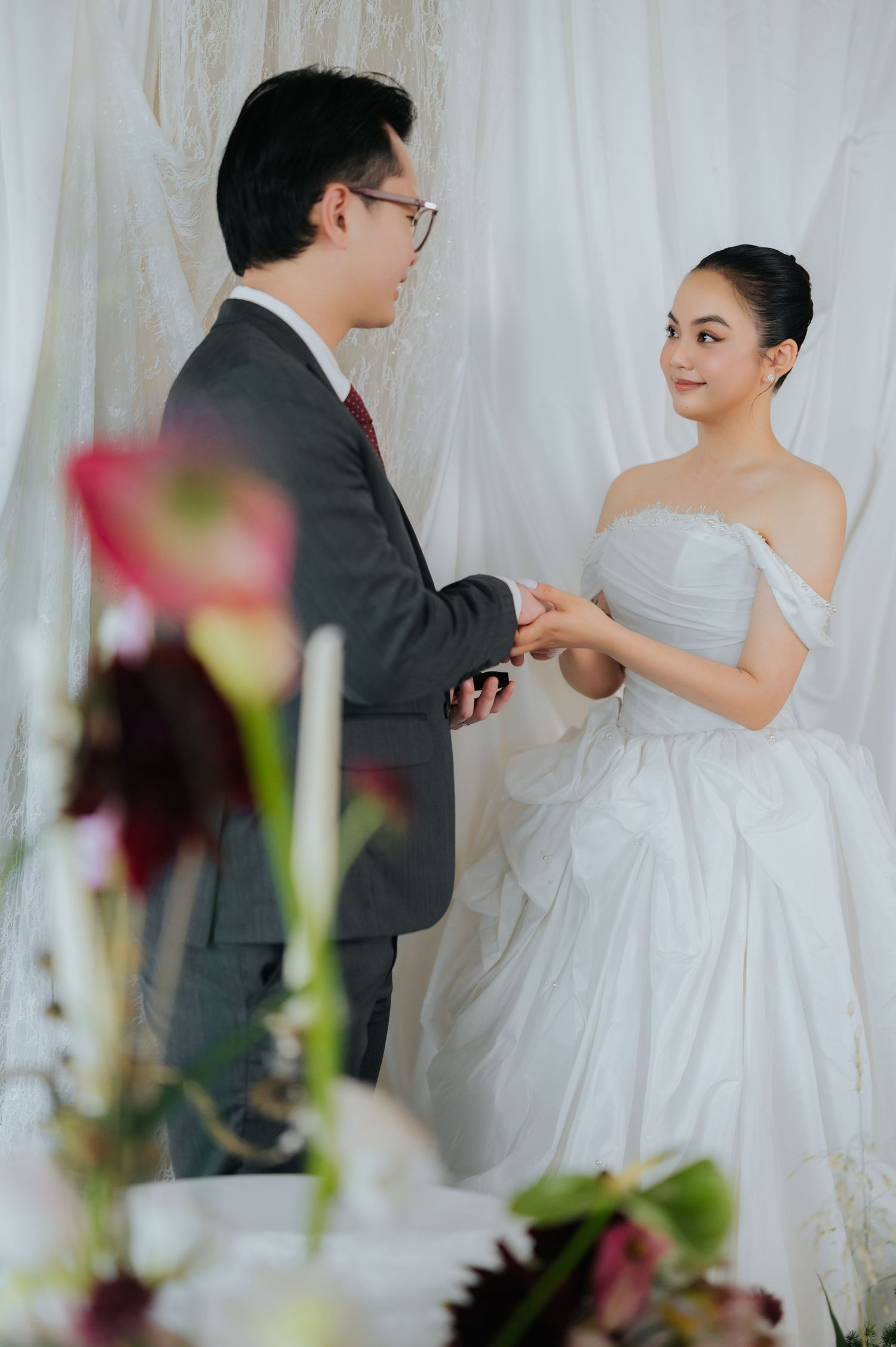 A groom in a gray suit exchanges rings with a bride in a white off-shoulder gown during an elegant wedding ceremony.