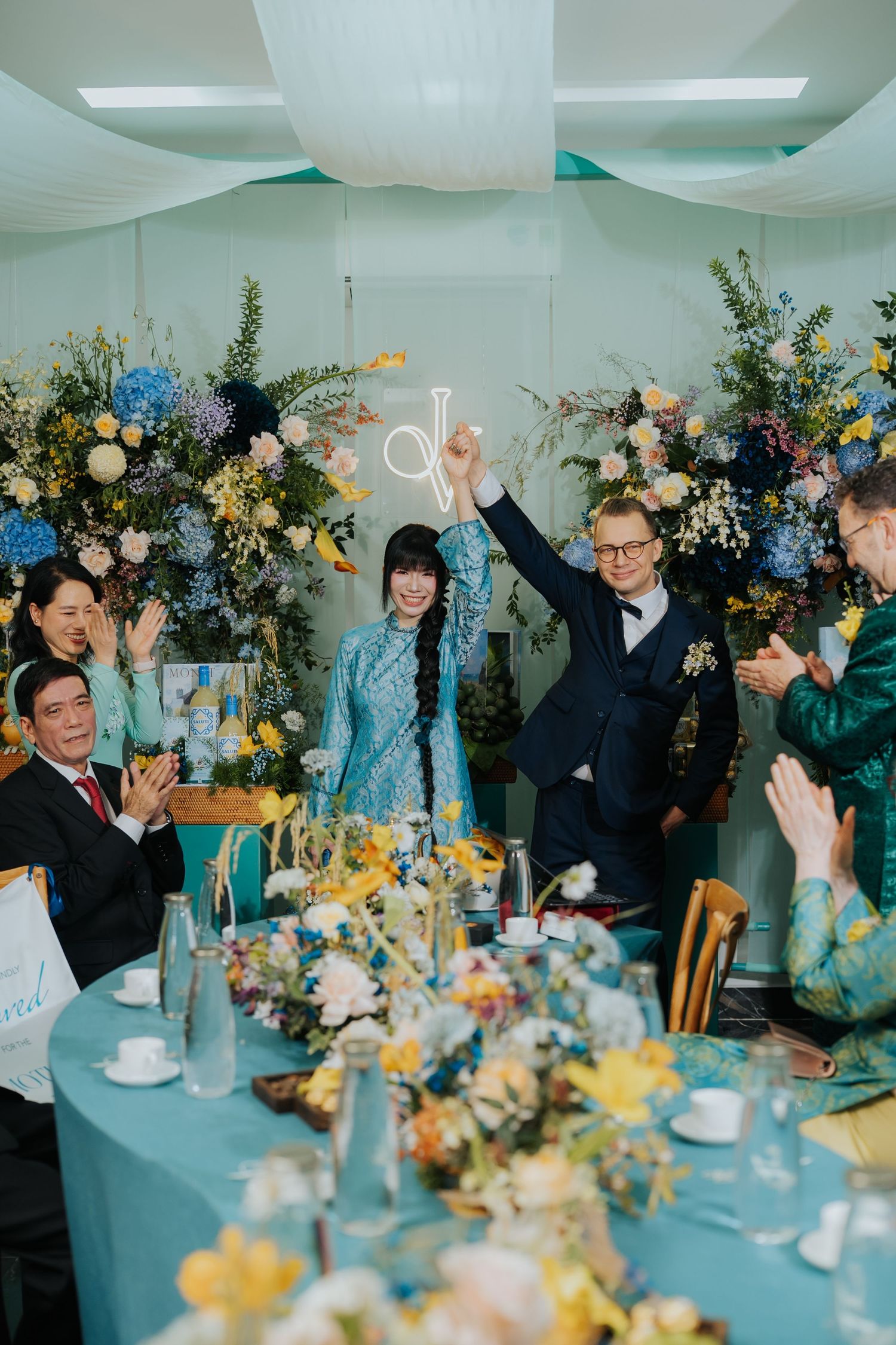 Joyful couple raises sparklers at a floral-decorated wedding reception with guests clapping around a teal table.