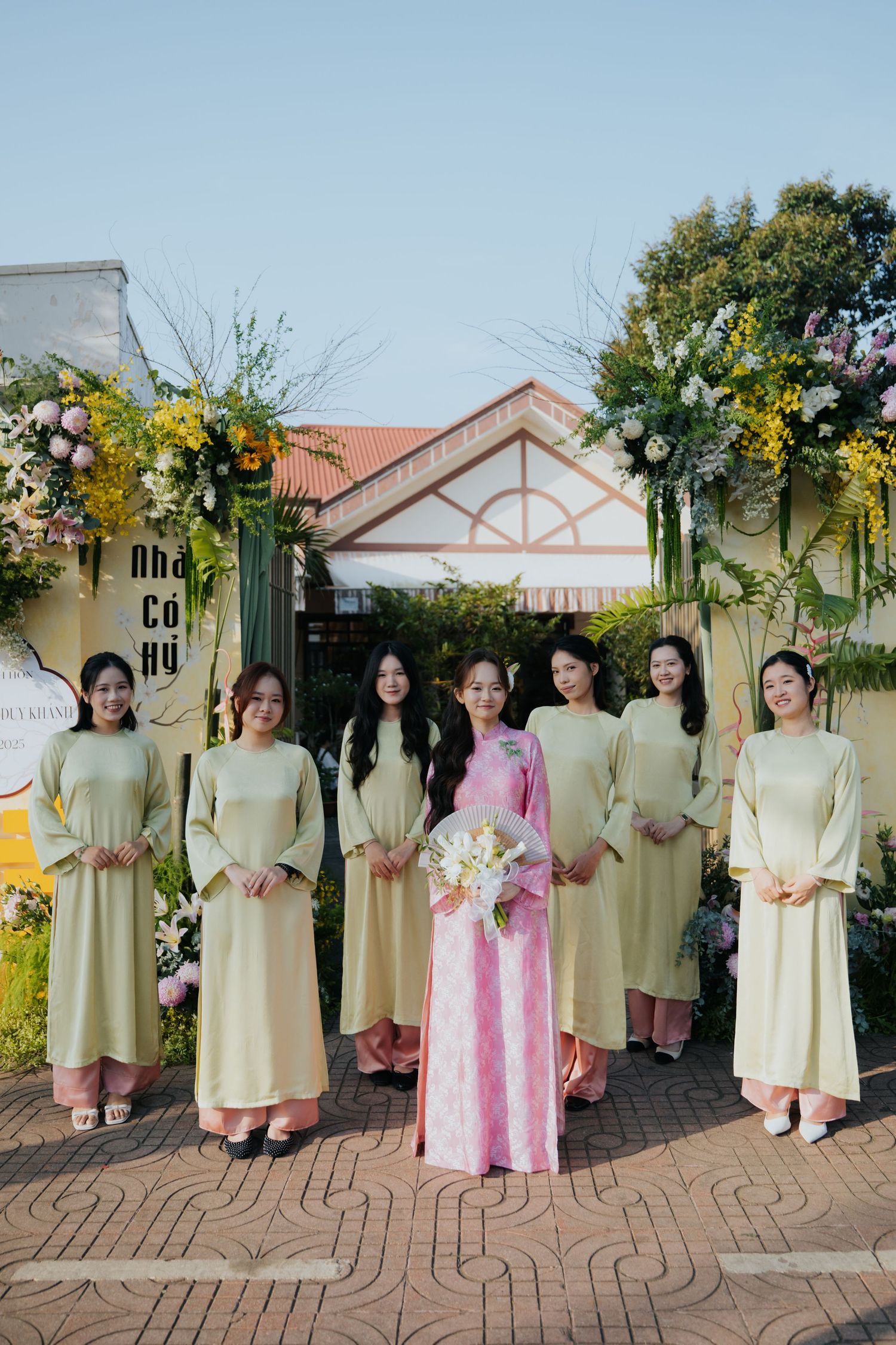 Bride in pink ao dai poses with bridesmaids in light yellow ao dai gowns at a Vietnamese wedding ceremony.