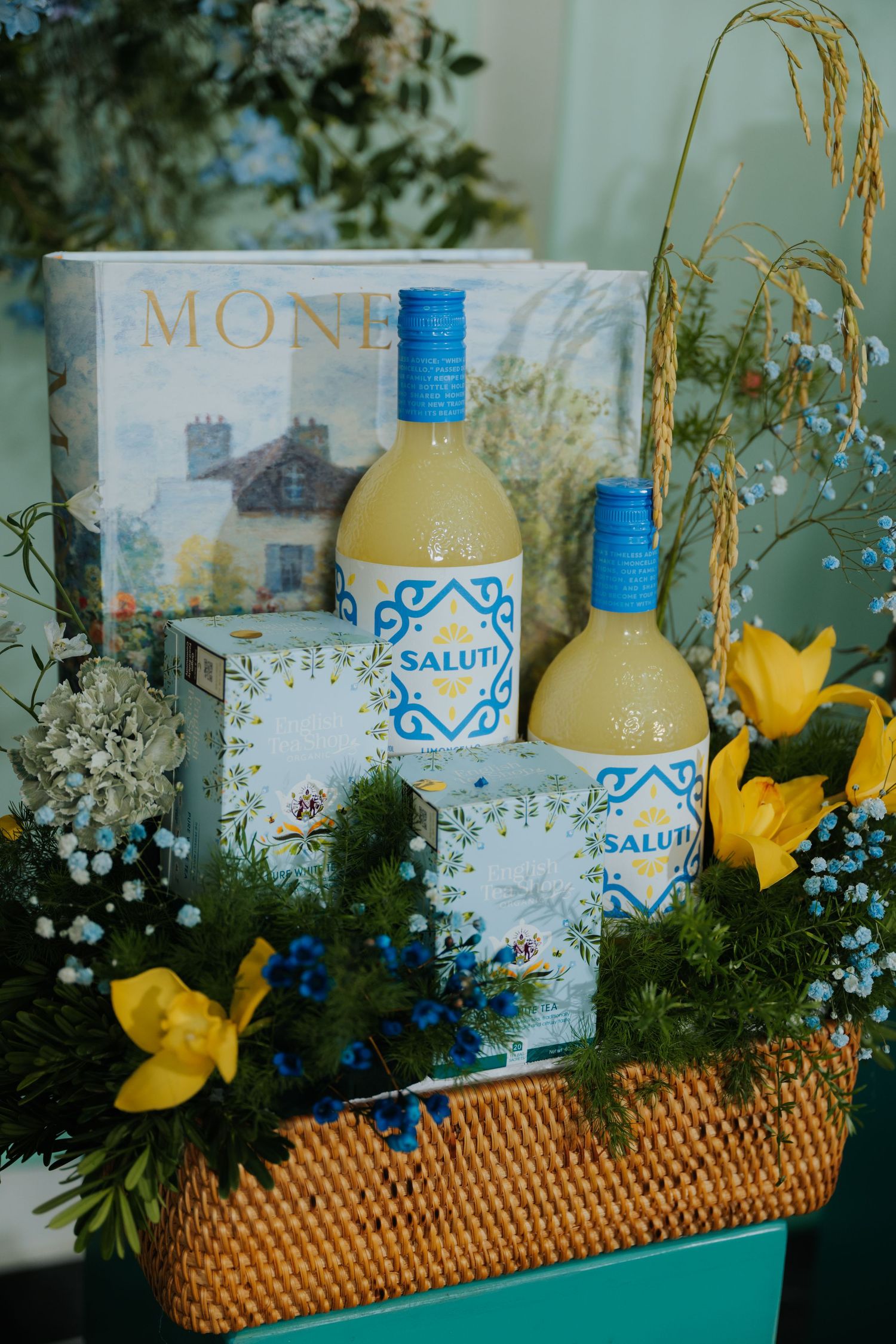 A gift basket with two Saluti bottles, small boxes, yellow flowers, and a Monet book arranged beautifully.