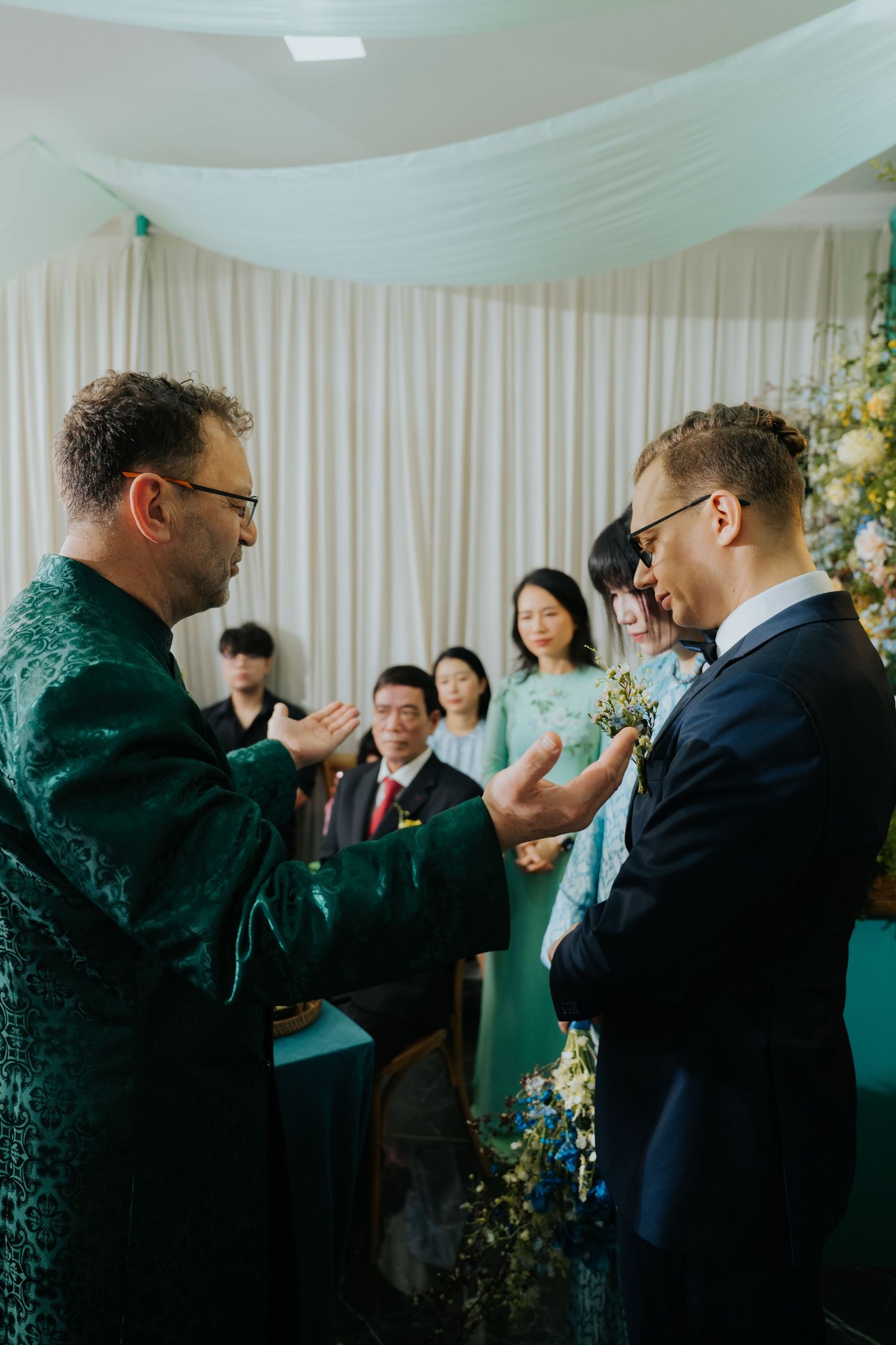 Two men in formal attire exchange words at a wedding ceremony with guests and floral arrangements in the background.
