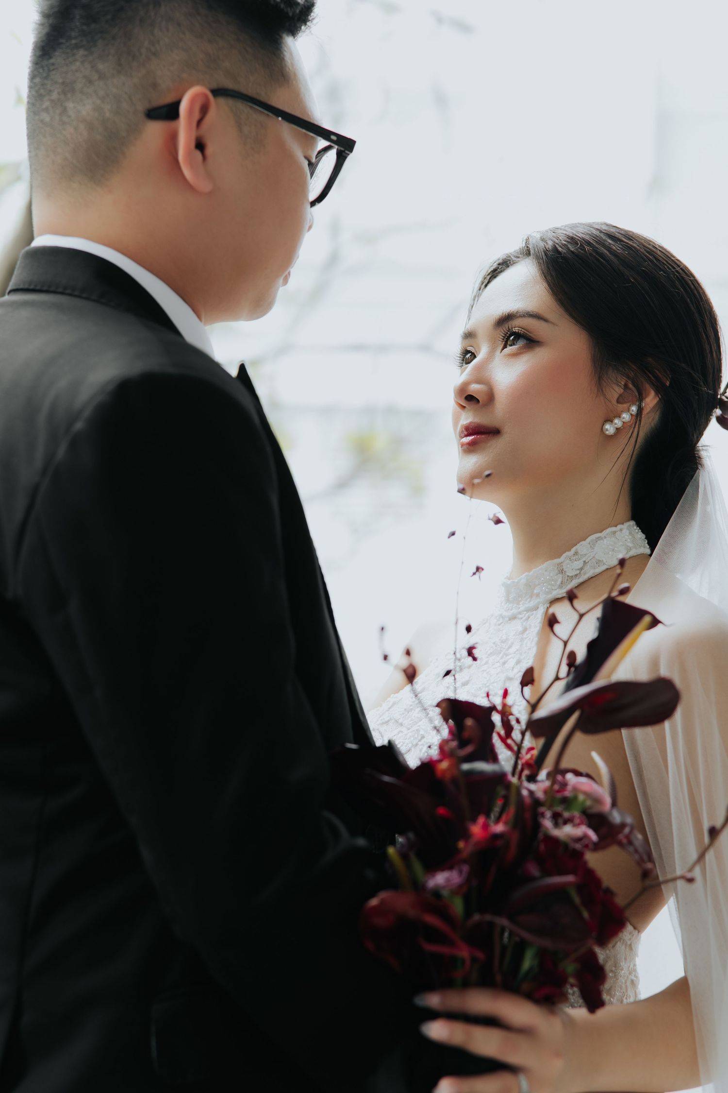 A bride holding a dark floral bouquet gazes lovingly at her groom in a black suit during their wedding ceremony.