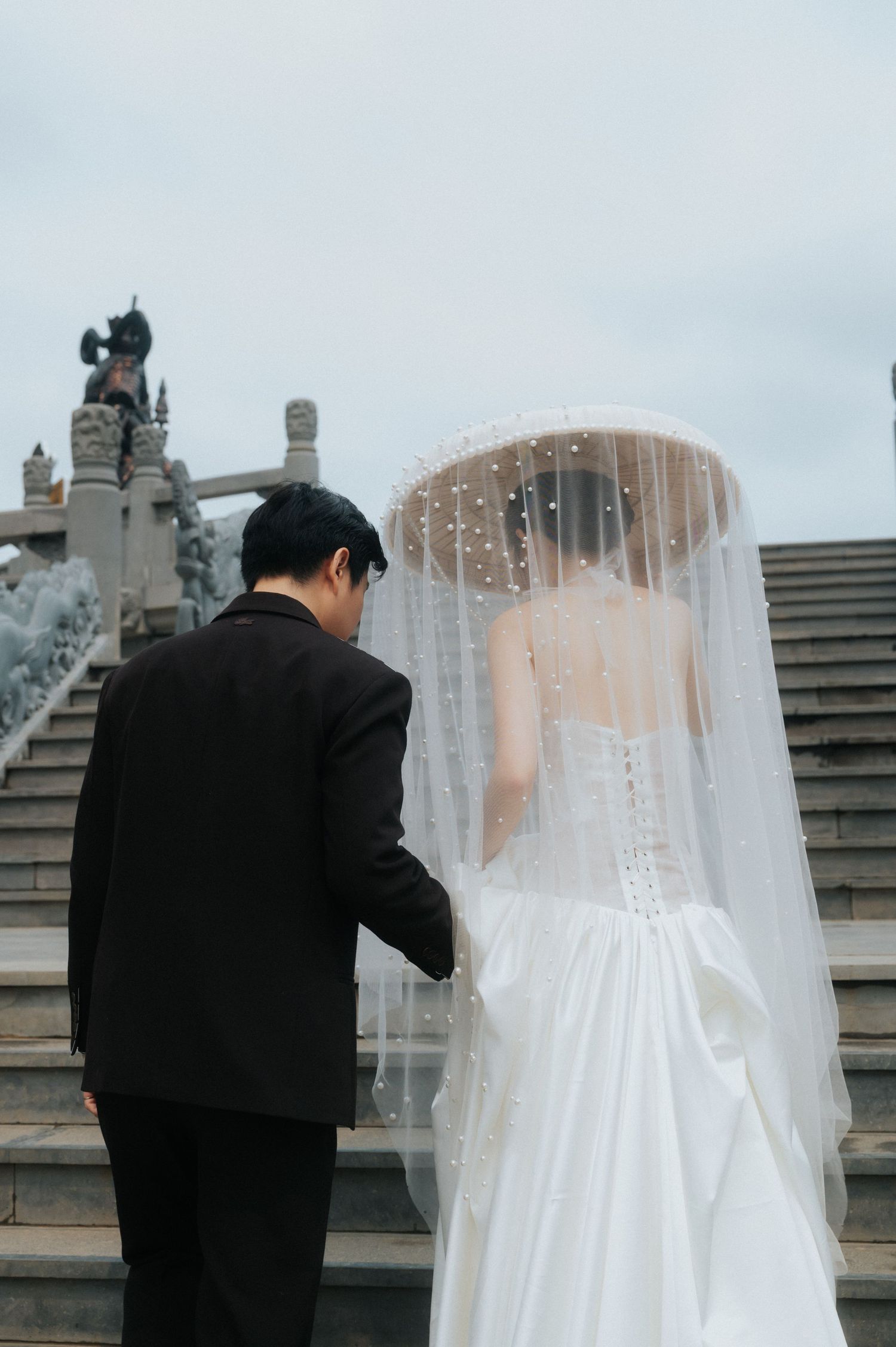 A couple in wedding attire holds hands ascending stone stairs, bride in white gown with pearl veil, groom in black suit.