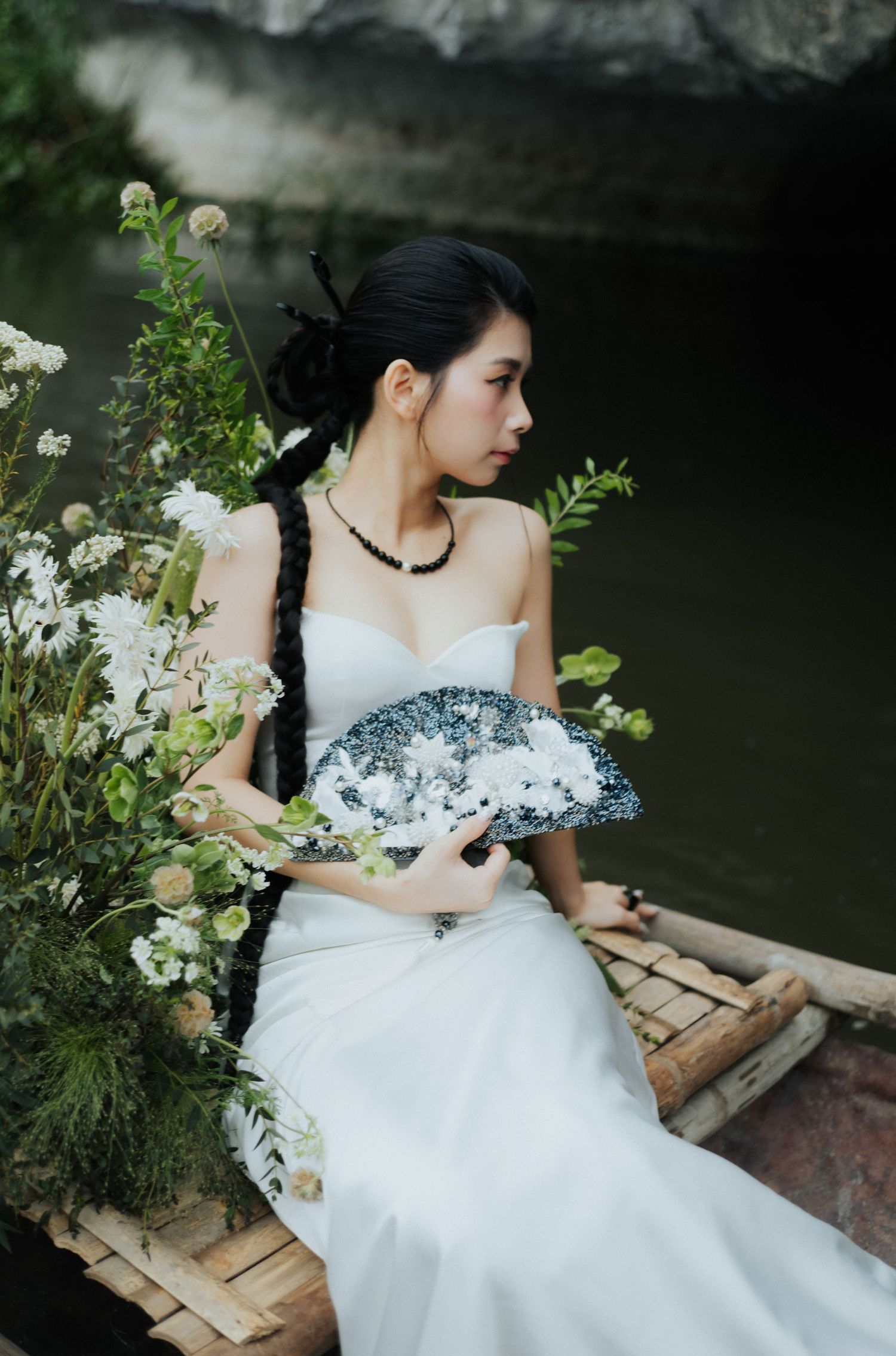 A bride in a white gown holds a floral fan, seated on bamboo near water surrounded by white blooms.