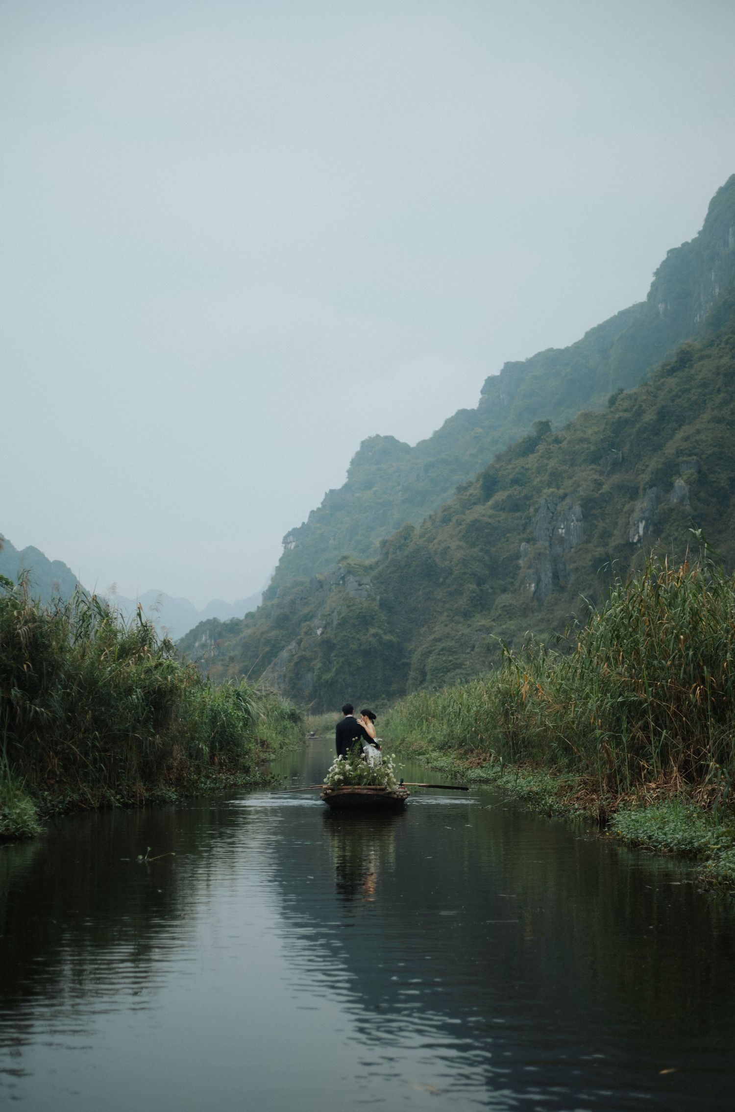 A lone boatman navigates a serene river through lush green karst mountains in misty Ninh Binh, Vietnam.