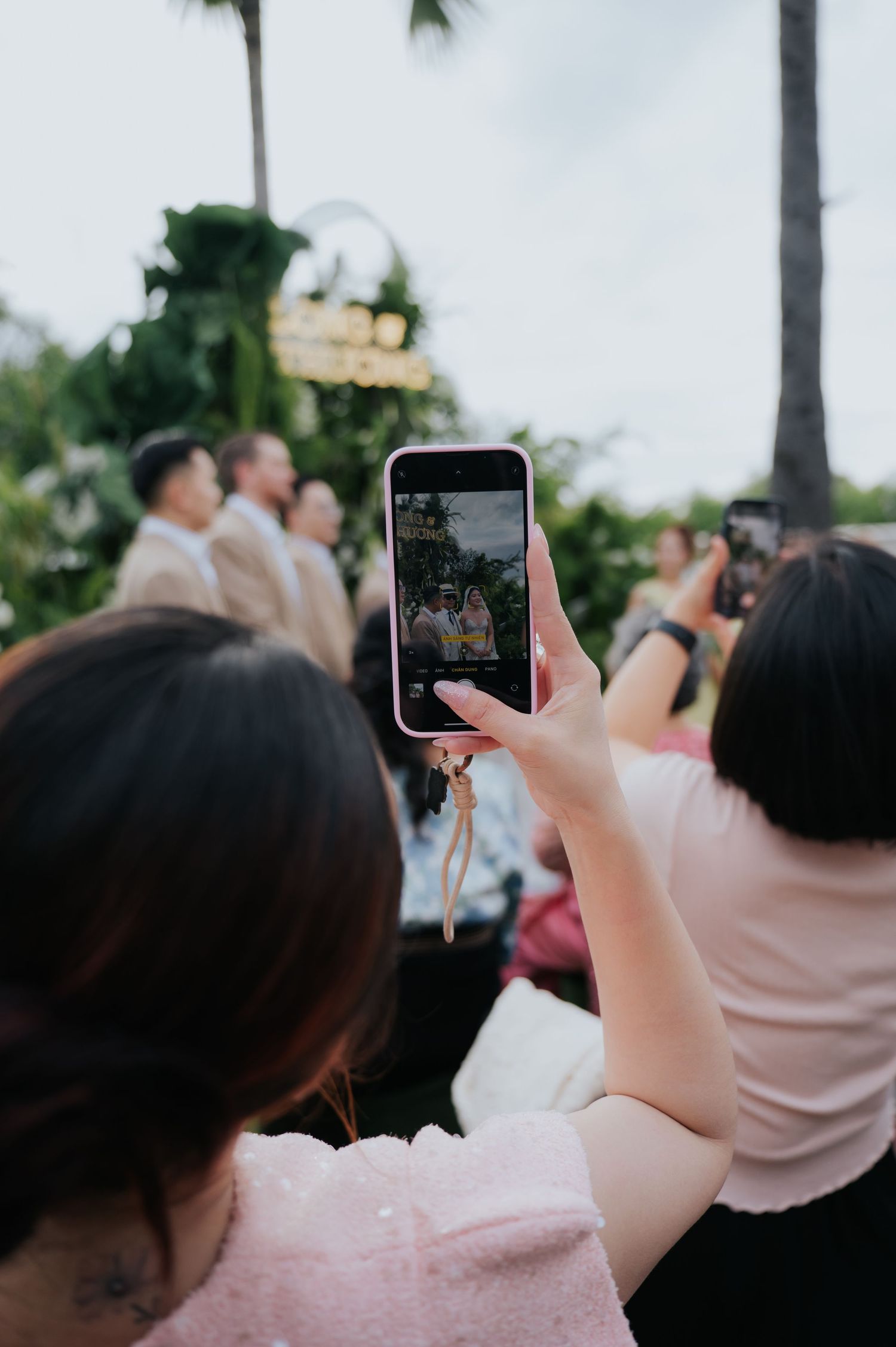 Guest capturing wedding ceremony moments on smartphone at outdoor tropical venue with palm trees.