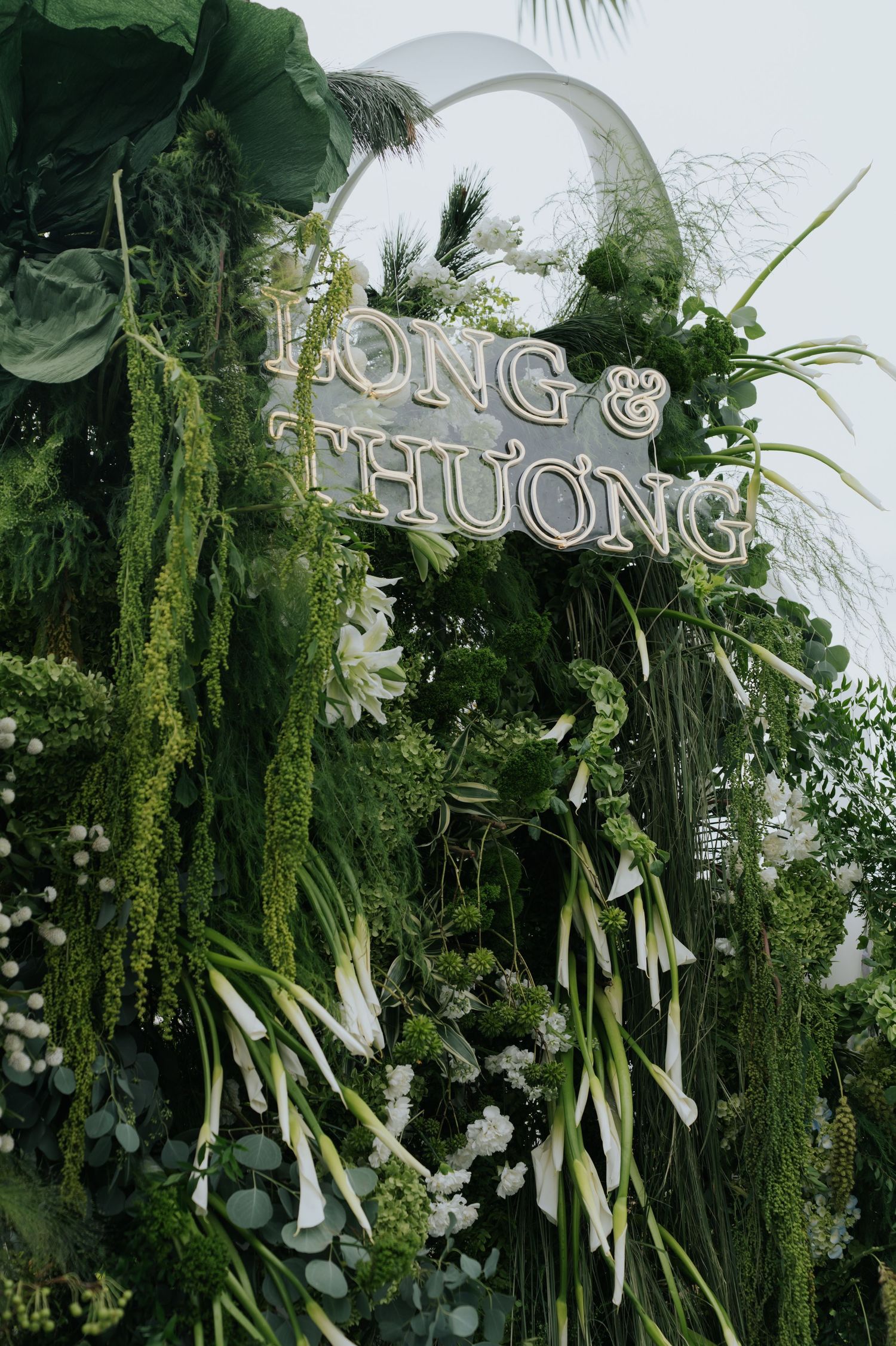 Lush green floral wedding backdrop with white calla lilies and a decorative sign reading Long and Thuong.