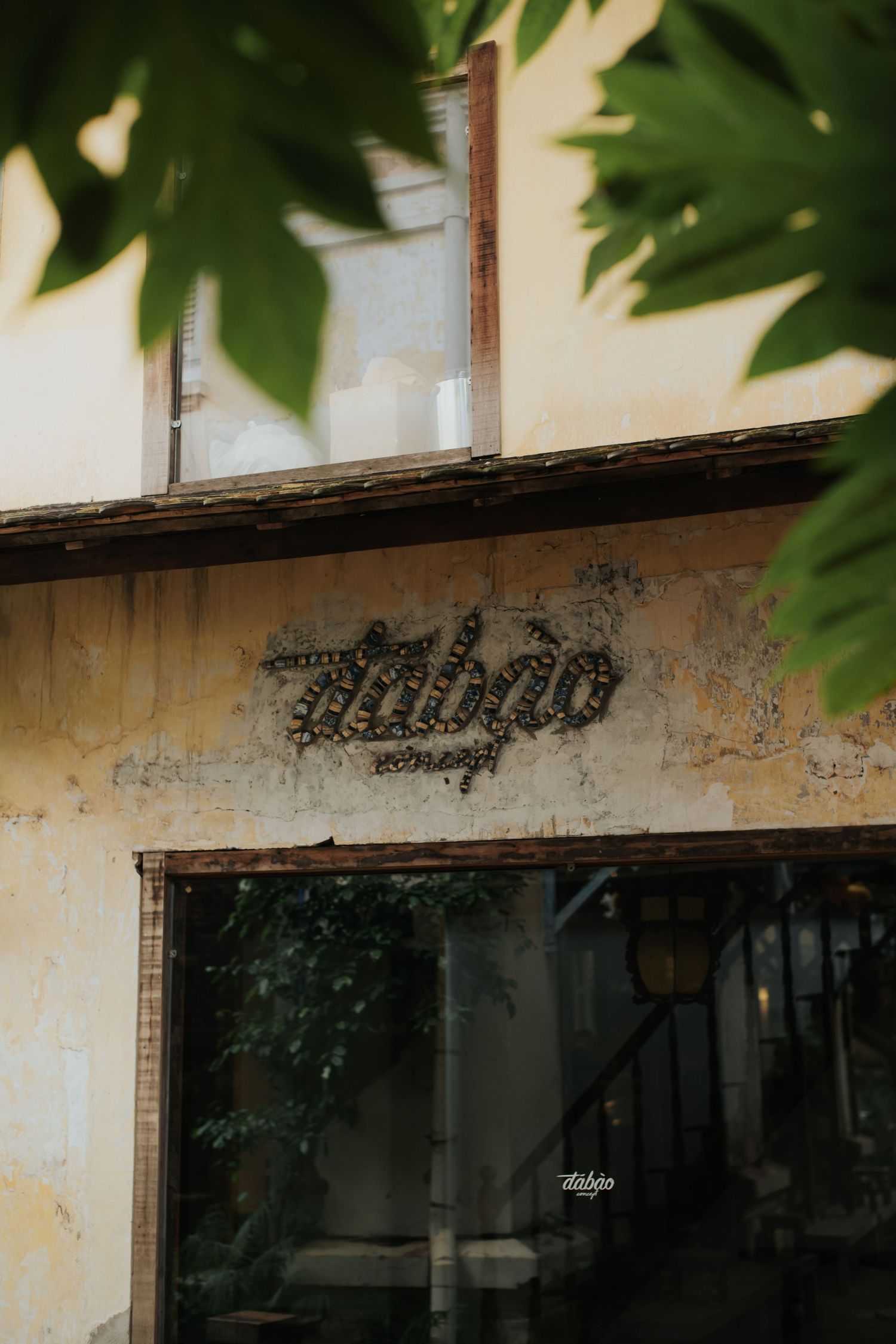Vintage weathered building facade with aged signage reading Tabao, framed by green leaves in soft natural light.