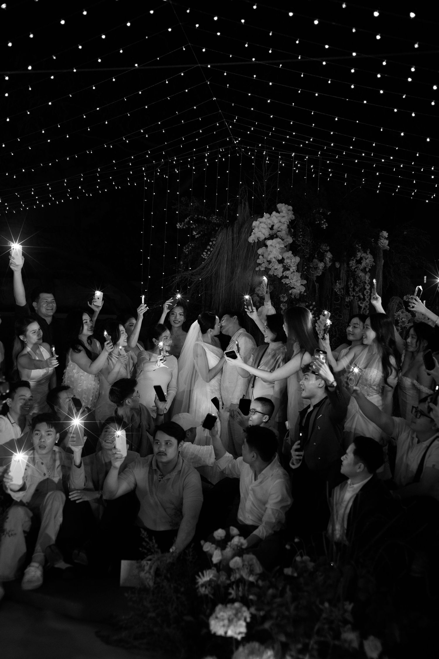 Black and white photo of wedding guests holding up phones with lights at a starlit indoor celebration.