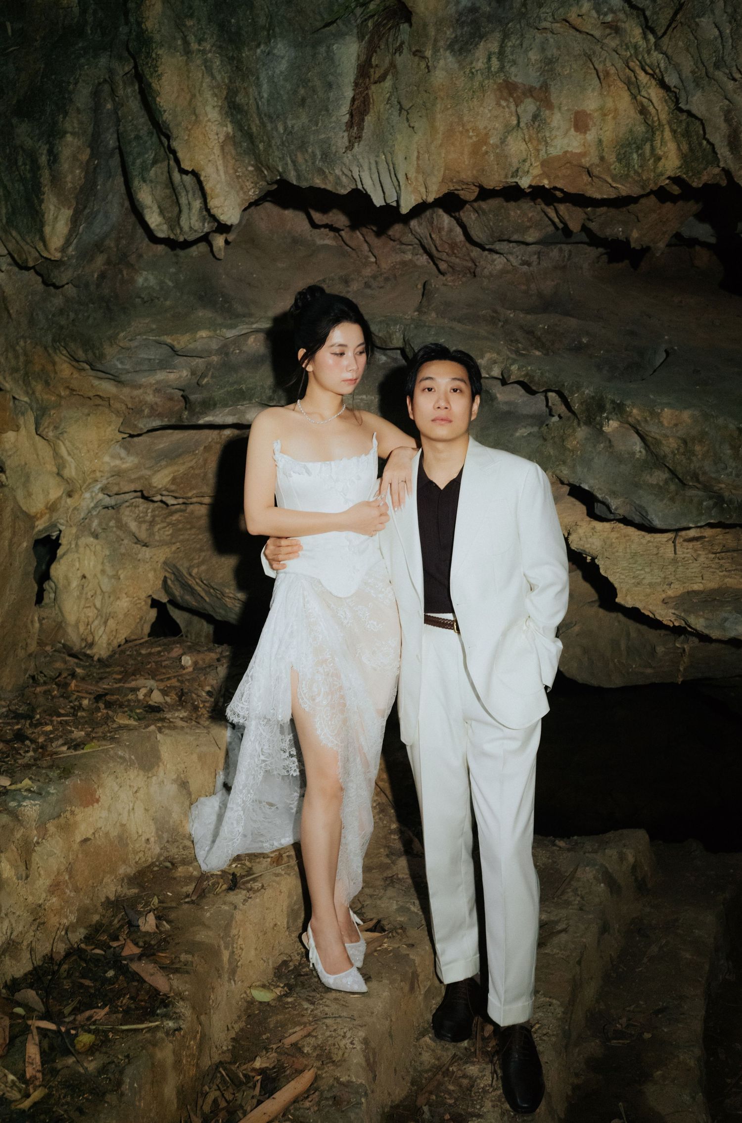Couple in white wedding attire posing romantically inside a dramatic cave with rocky walls.