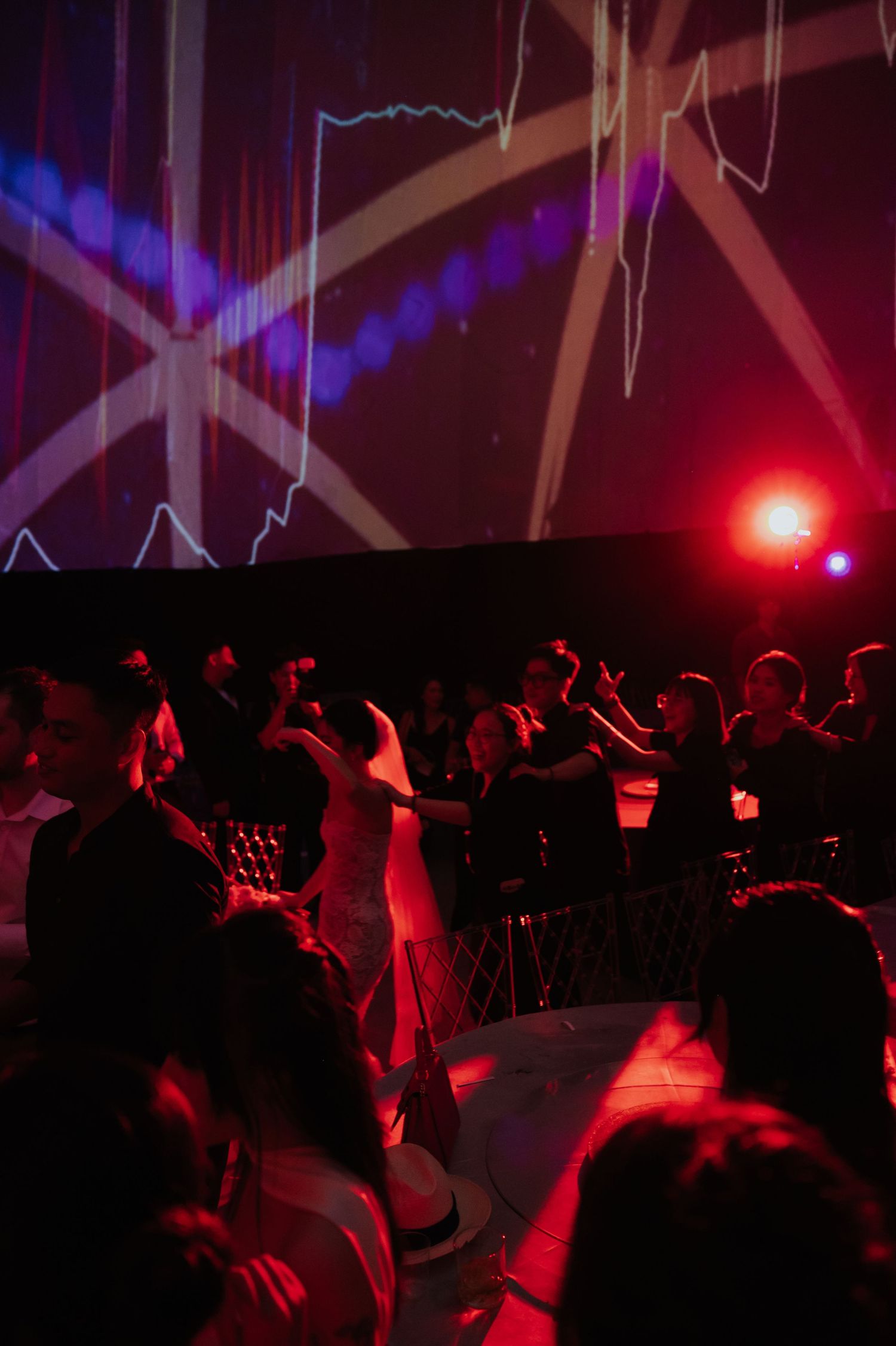Guests dancing at a wedding reception under dramatic red and purple stage lighting with projected visuals.