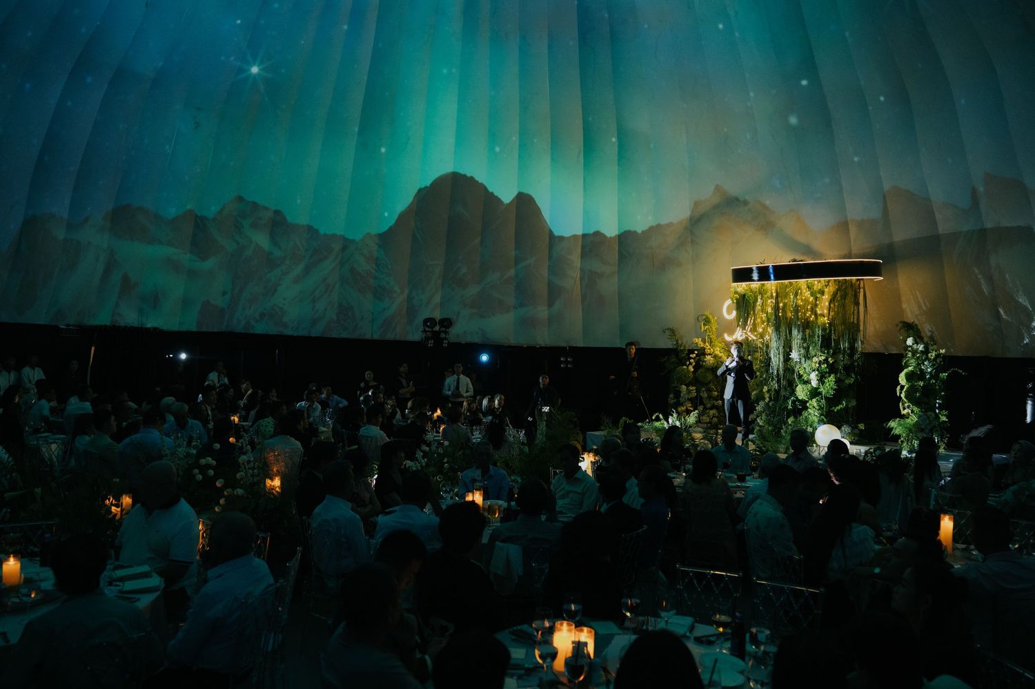 Elegant gala dinner under aurora borealis projection in a tent venue with candlelit tables and a speaker on stage.
