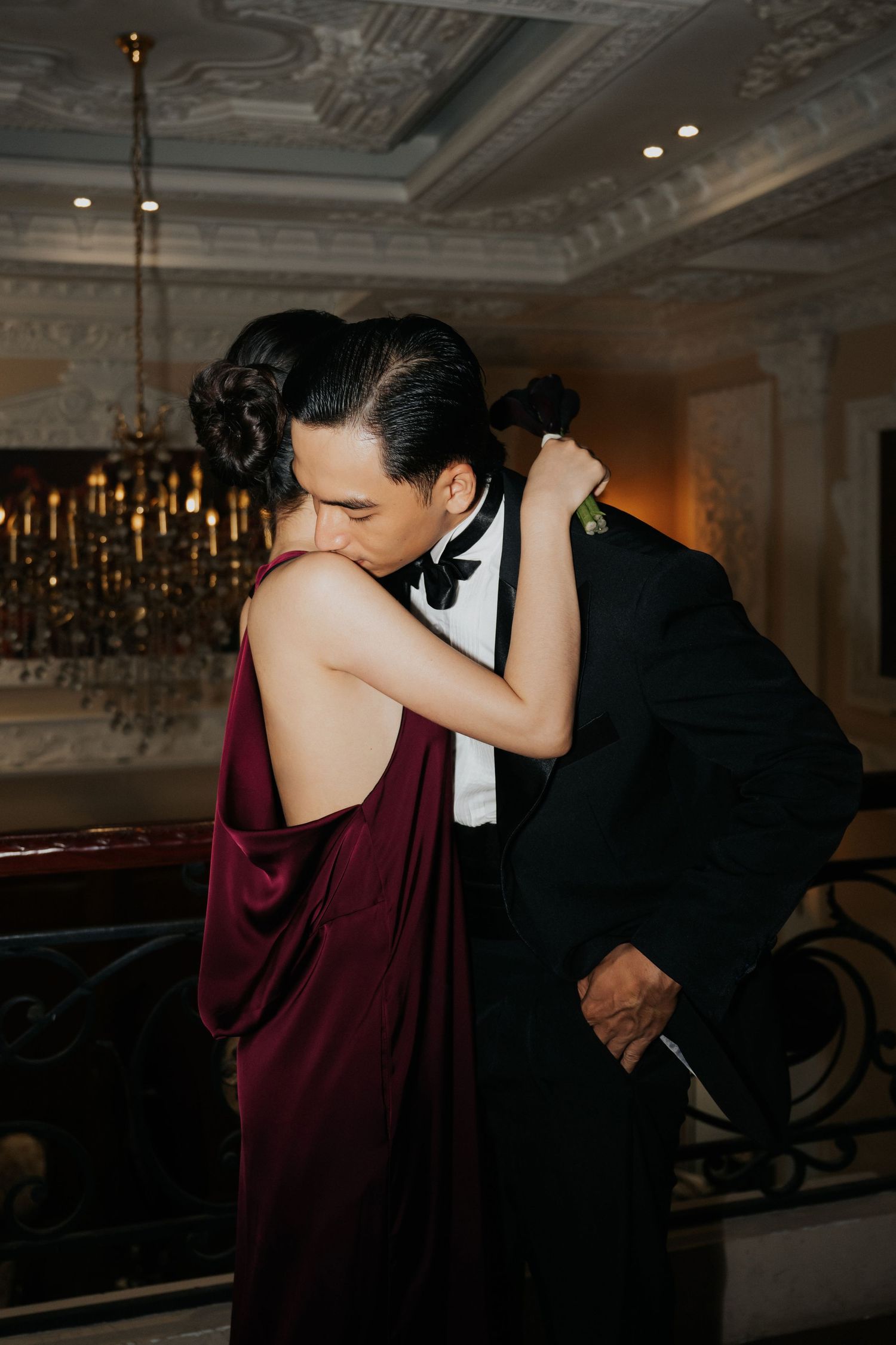 Couple embracing in elegant ballroom, woman in burgundy backless gown, man in black tuxedo, ornate chandelier backdrop.