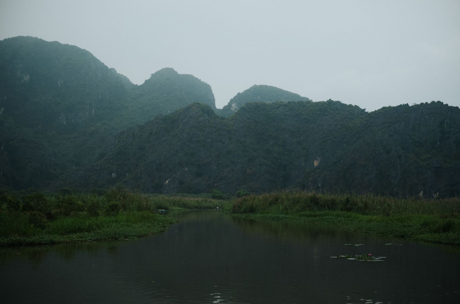 Misty karst mountains reflected in a calm river surrounded by lush green vegetation in Ninh Binh, Vietnam.