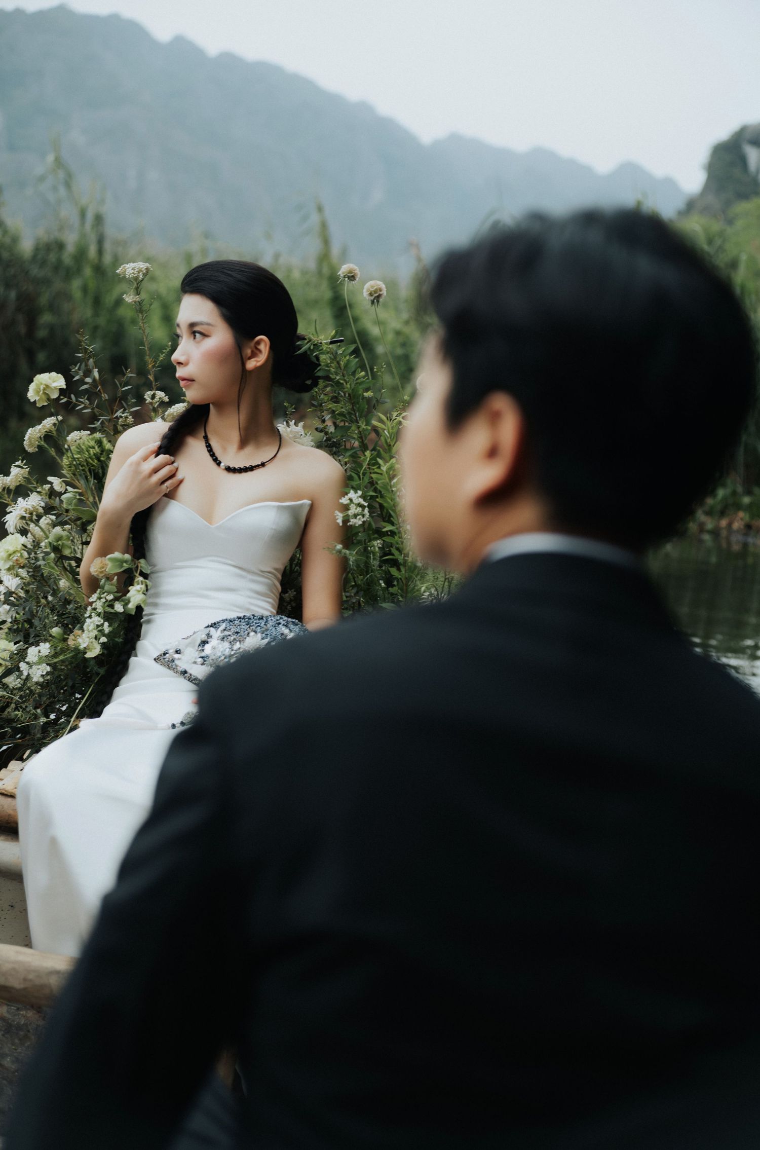 A bride in a white strapless gown sits among wildflowers as a groom in a black suit sits in the foreground.