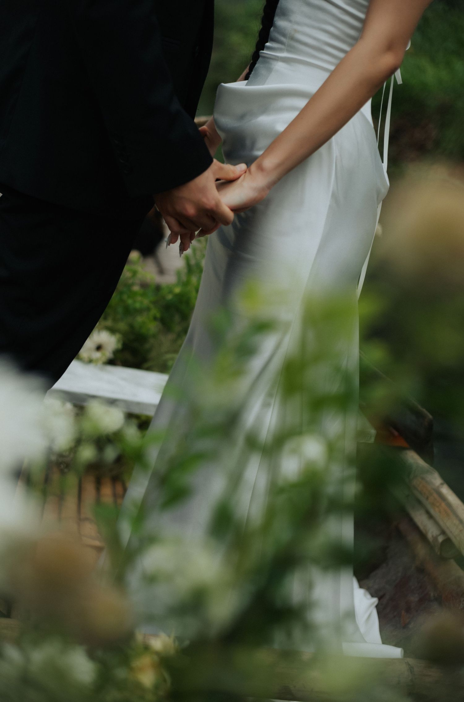Bride and groom holding hands outdoors during wedding ceremony, surrounded by white flowers and lush greenery.