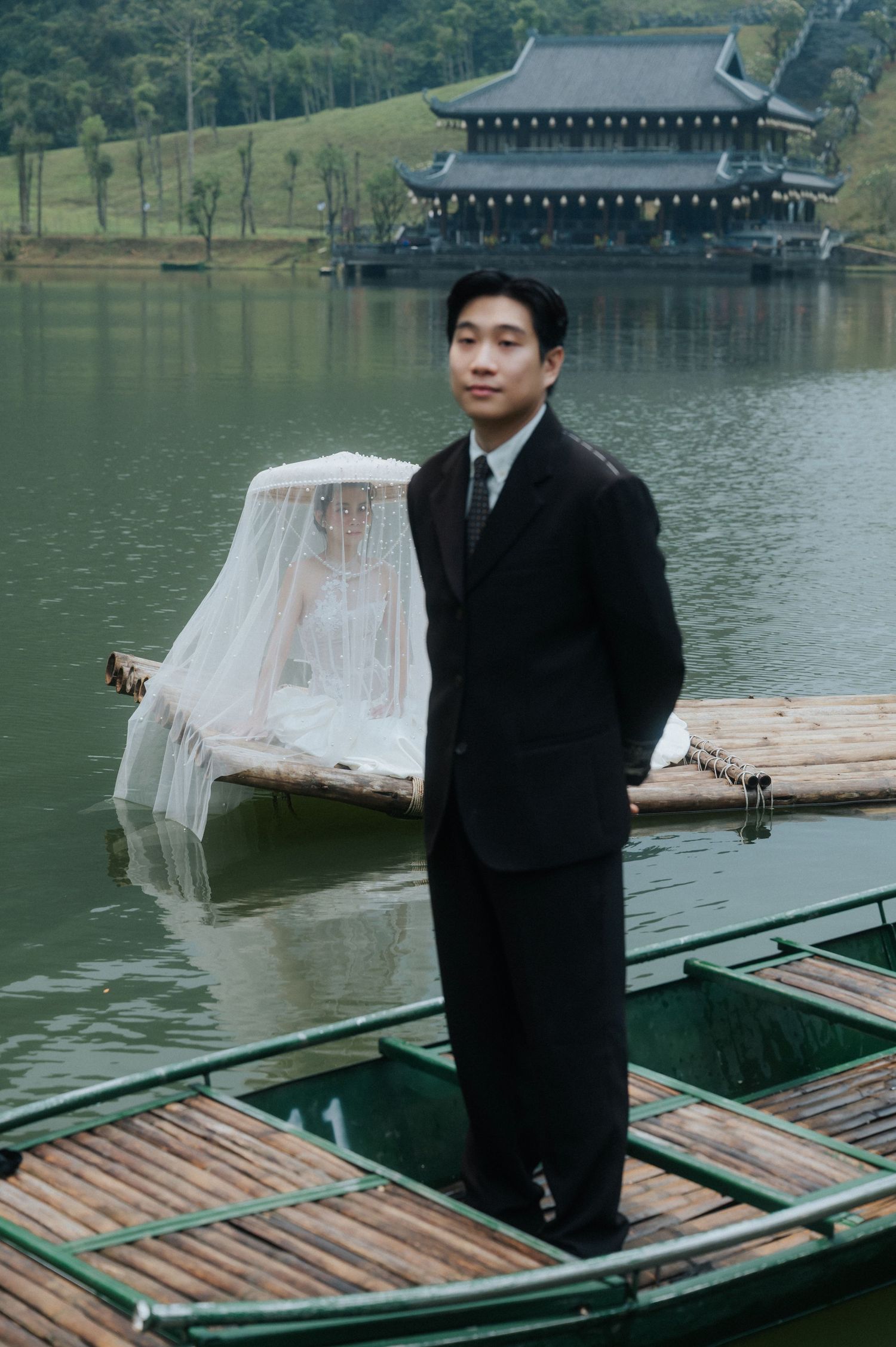 A groom in a black suit stands on a bamboo raft beside a veiled bride near a lakeside pagoda.