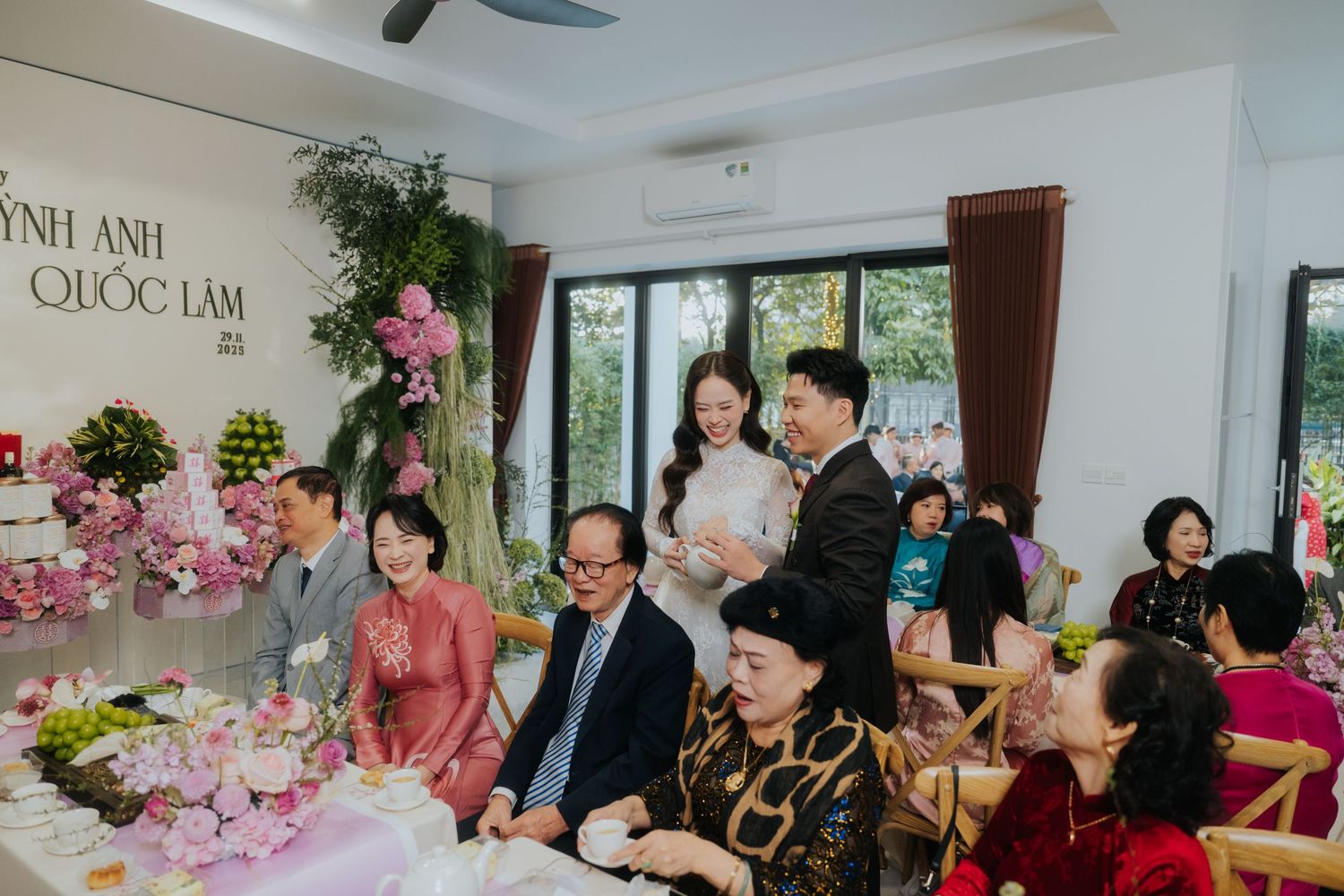 Vietnamese wedding ceremony with guests in traditional ao dai attire gathered around decorated tables with pink floral arrangements.