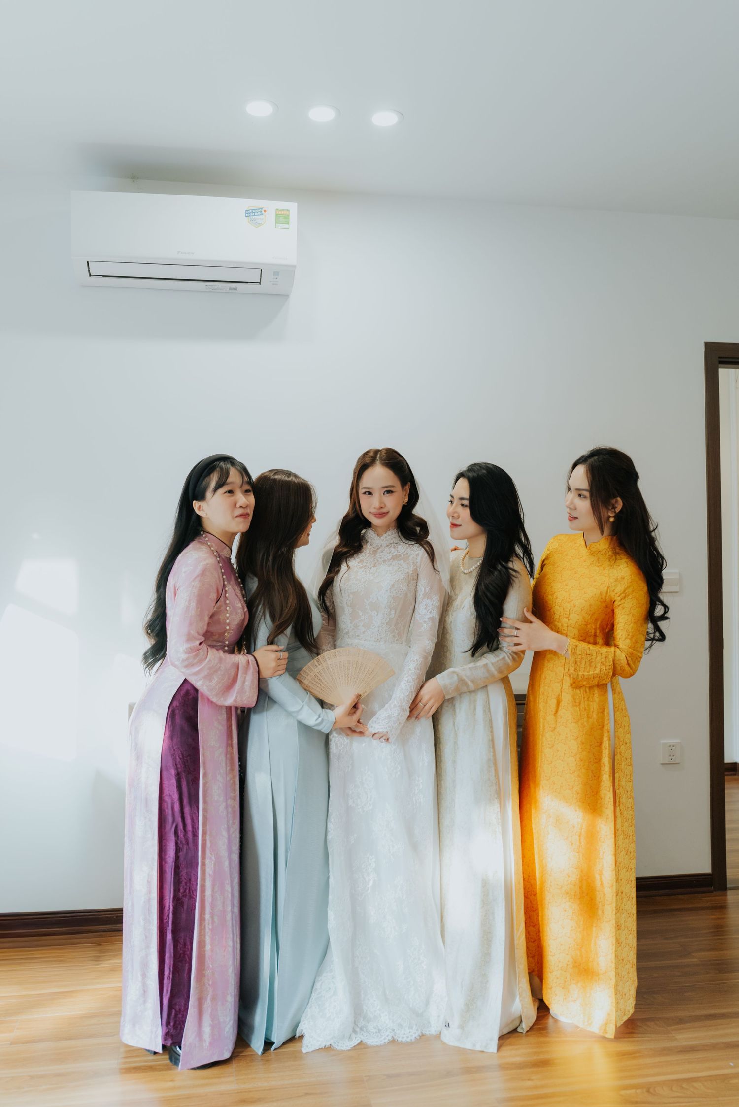 Five women in elegant Vietnamese ao dai dresses pose together indoors, with bride in white lace gown surrounded by colorful bridesmaids.