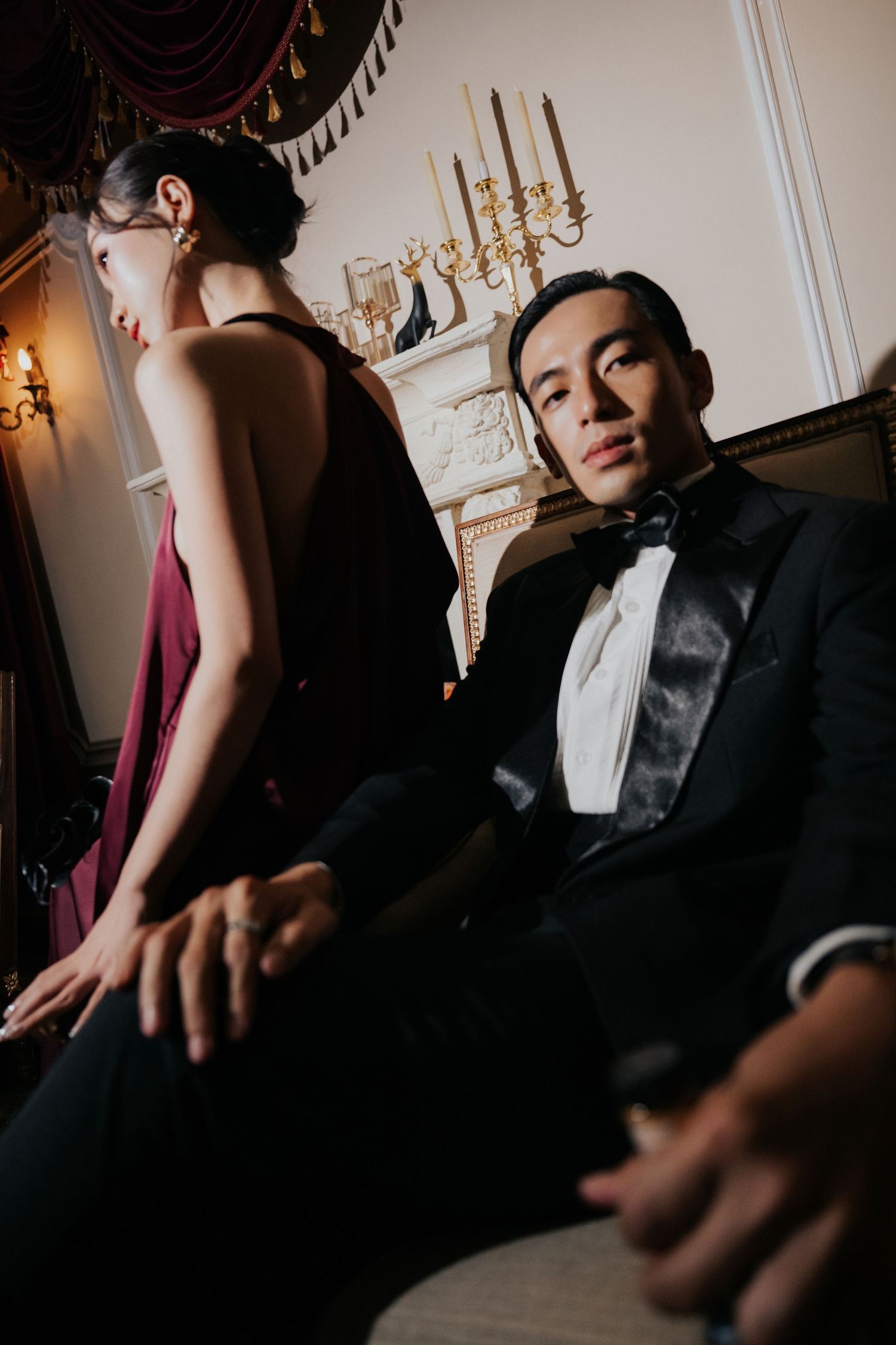 A man in a tuxedo sits elegantly beside a woman in a burgundy gown in a luxurious candlelit room.
