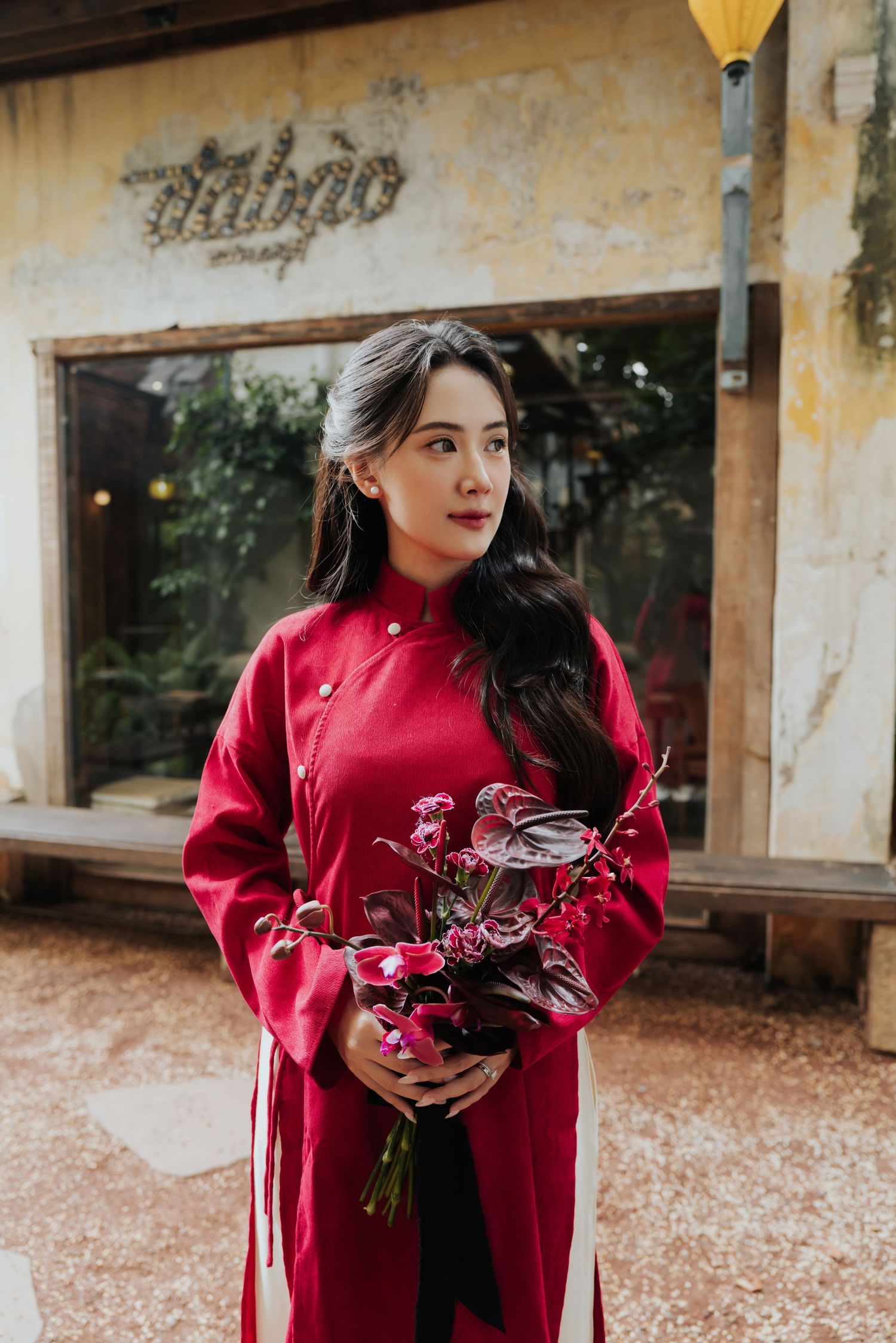 A young woman in a red ao dai holds a bouquet of red flowers, standing before a rustic building in Vietnam.