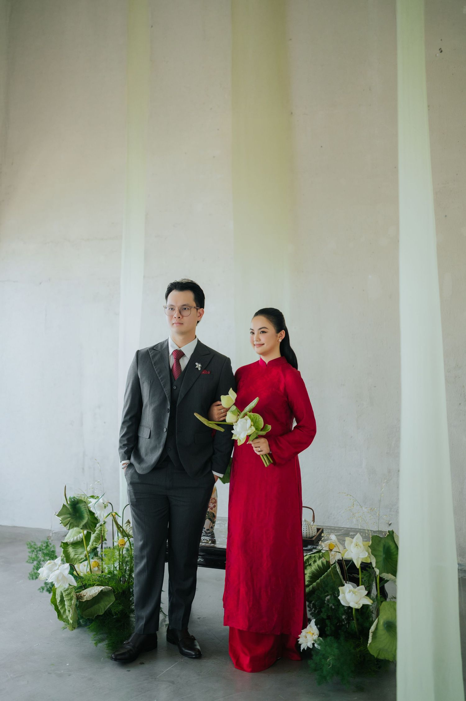 Couple in traditional Vietnamese wedding attire, man in gray suit and woman in red ao dai holding white flowers.