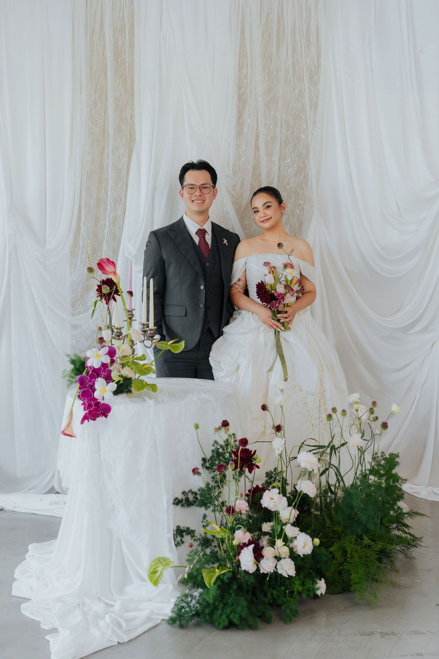 Bride in white gown and groom in gray suit pose by floral arrangements with white drape backdrop.