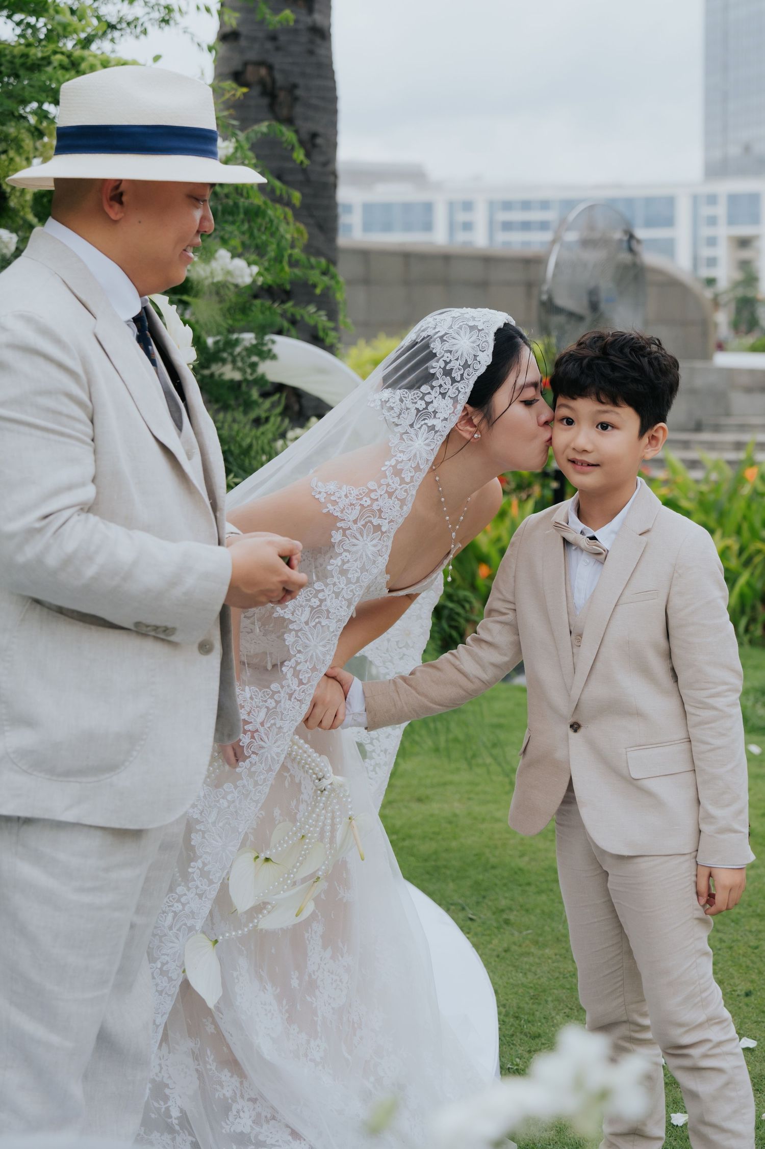Bride in white lace gown kisses young boy in beige suit at outdoor wedding ceremony with groom in white suit nearby.