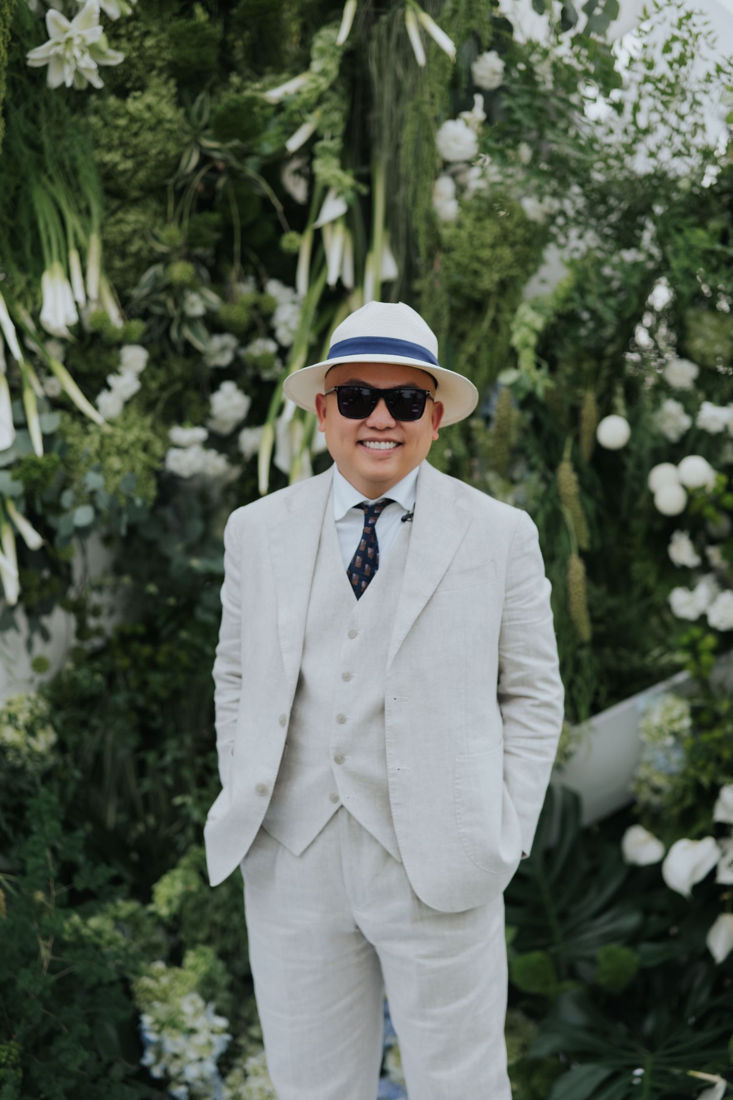 Man in white linen suit, panama hat, and sunglasses smiling at a floral wedding backdrop.