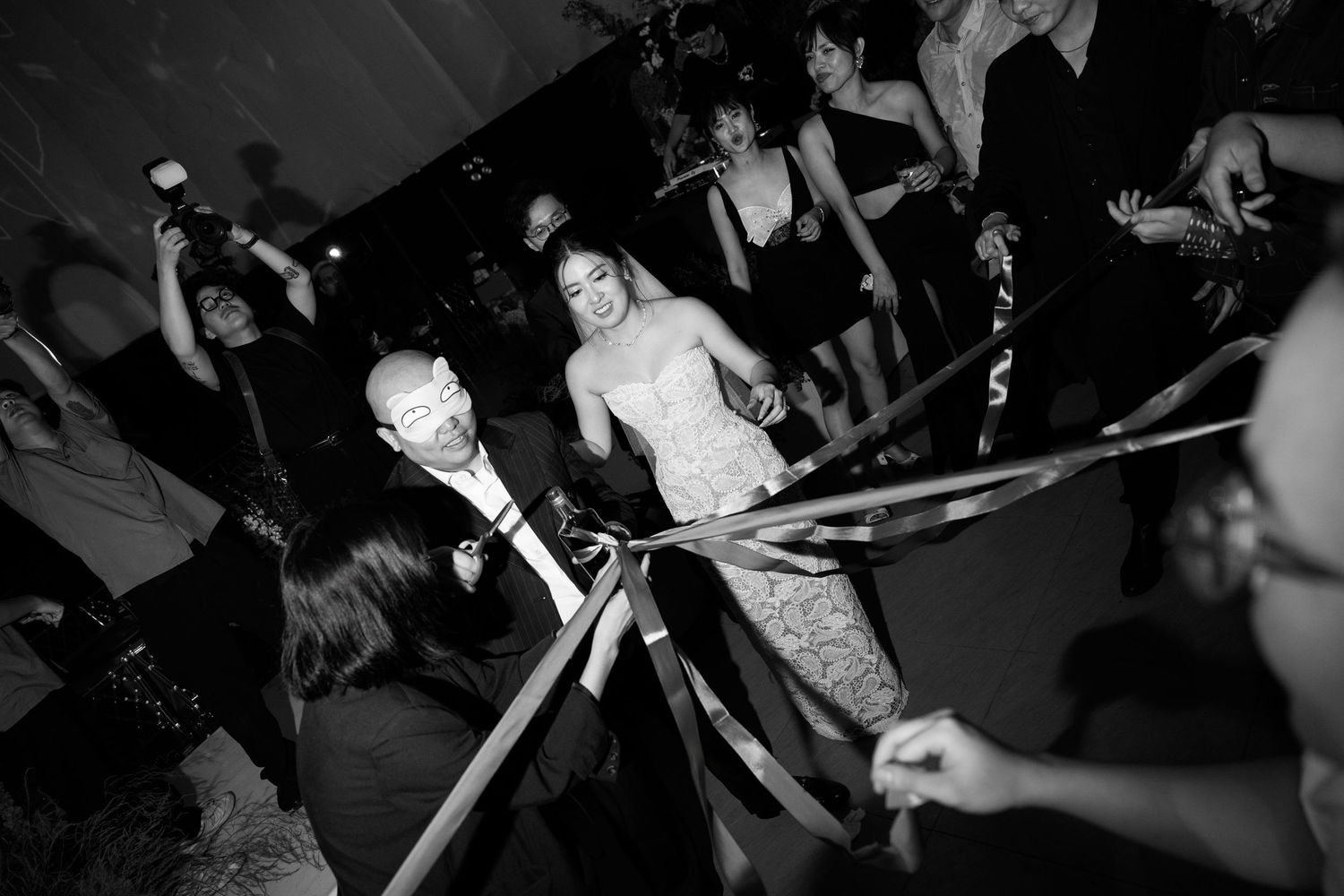 Black and white photo of elegantly dressed couple cutting ribbon at formal event surrounded by celebrating guests.