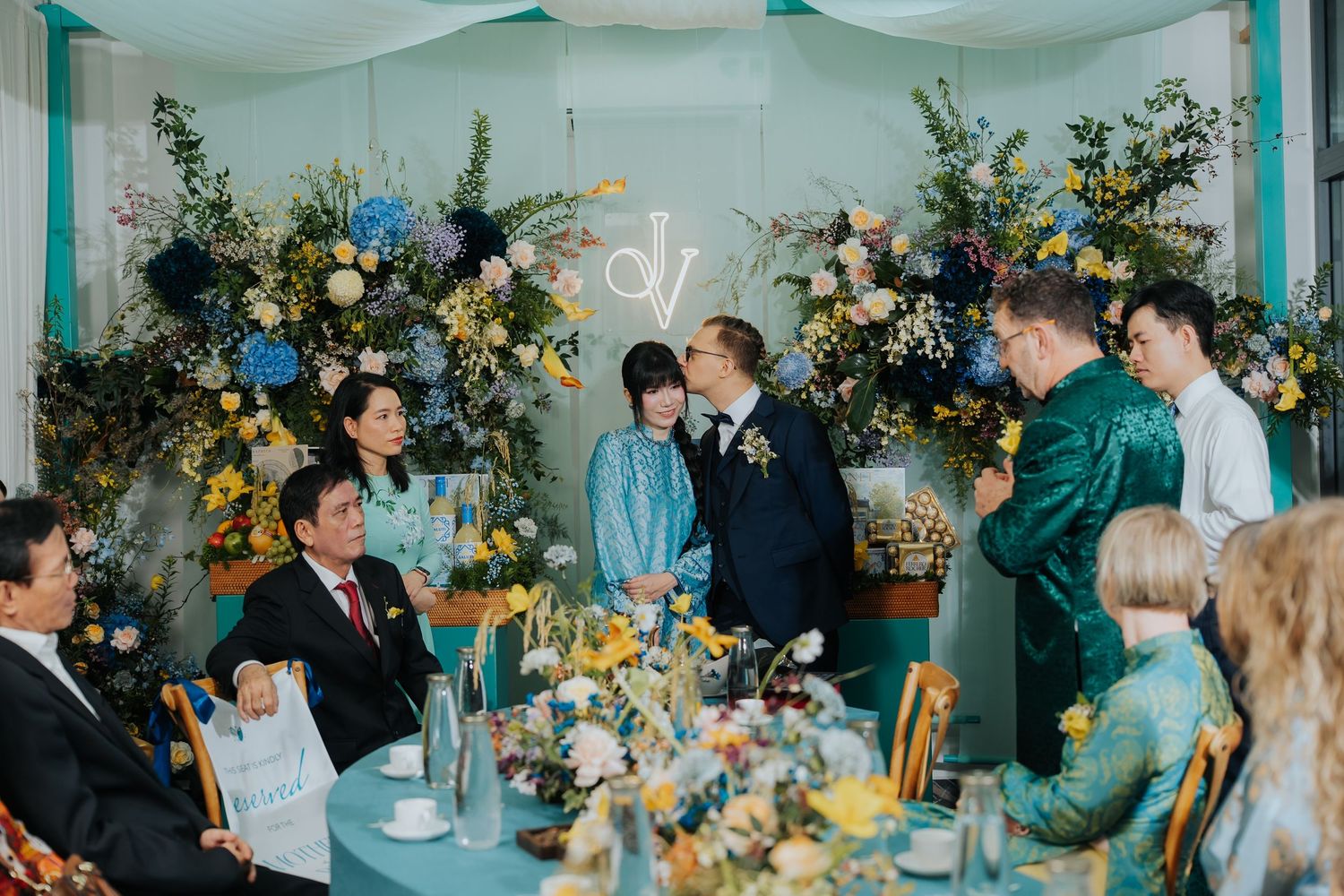 Wedding reception with guests gathered around a floral-decorated table, featuring blue and yellow arrangements.